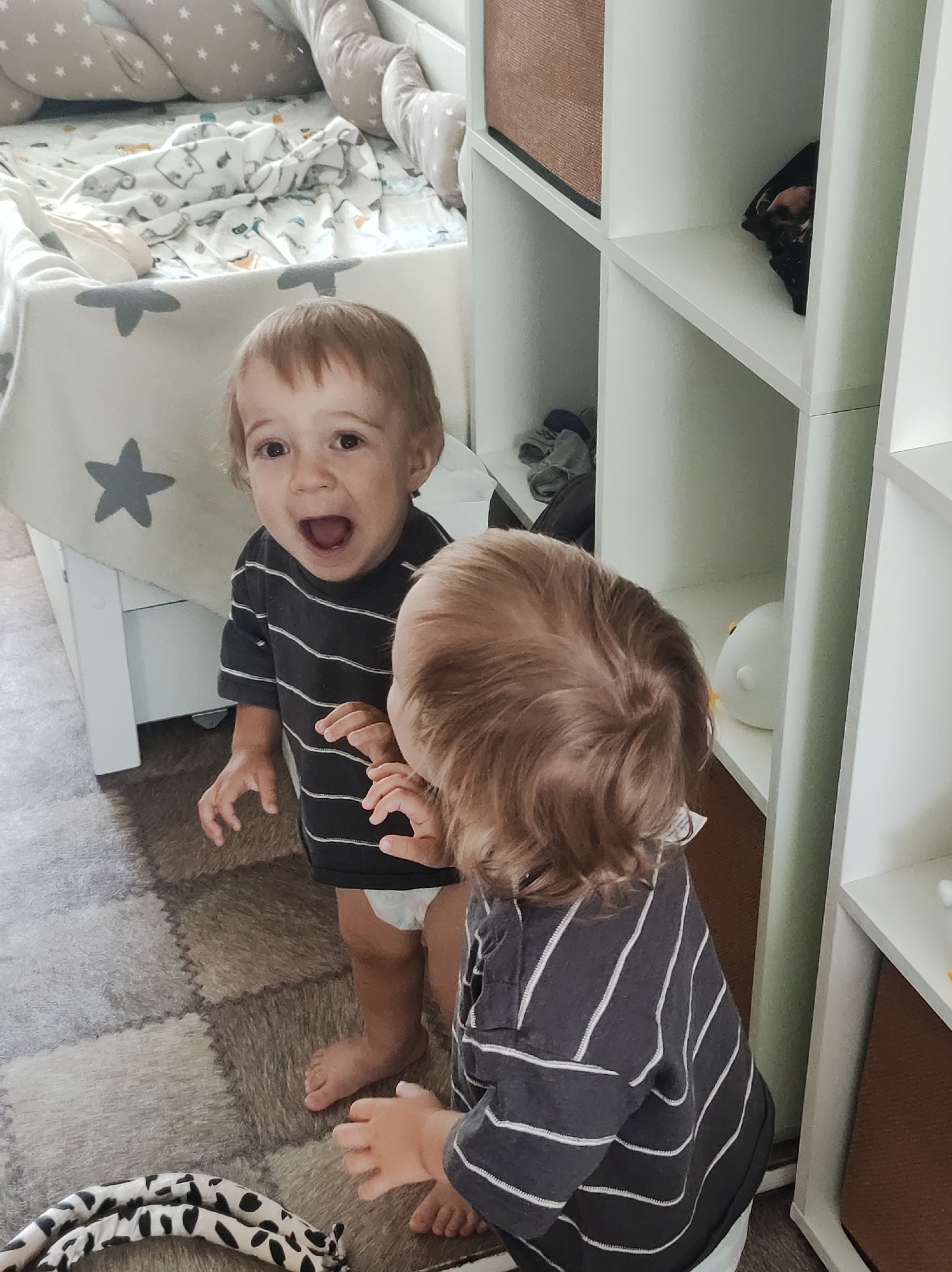 A toddler boy is standing in front of a mirror and looking at his reflection. He is wearing a dark gray striped t-shirt and a diaper. His mouth is open in a surprised expression. The boy is in a room with a white crib in the background and a white shelf unit to the right. The floor is covered with a patterned rug. The overall impression is one of playfulness and curiosity. This image is not located in Chaikovsky, Russia.