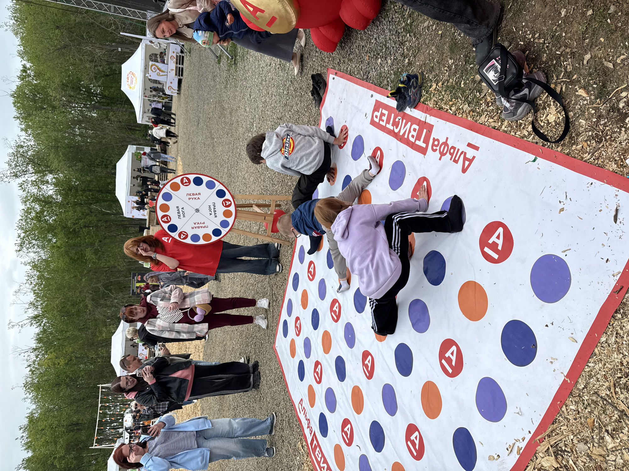 In Paratunka, Russia, an outdoor festival is underway with a game of Twister set up on a white mat with colorful circles. A group of people are engaged in the game, with several children and adults positioned on the mat in various poses. In the foreground, a person in a purple hoodie is on the mat, while a person in a gray hoodie is also playing. A younger child in a blue jacket is further back on the mat. A spinning wheel with color and body part indicators stands to the left of the mat, with a person in a red shirt near it. In the background, tents are set up, suggesting a fair or event. The ground is covered in gravel and wood chips. The overall atmosphere appears to be one of recreational activity and community gathering.