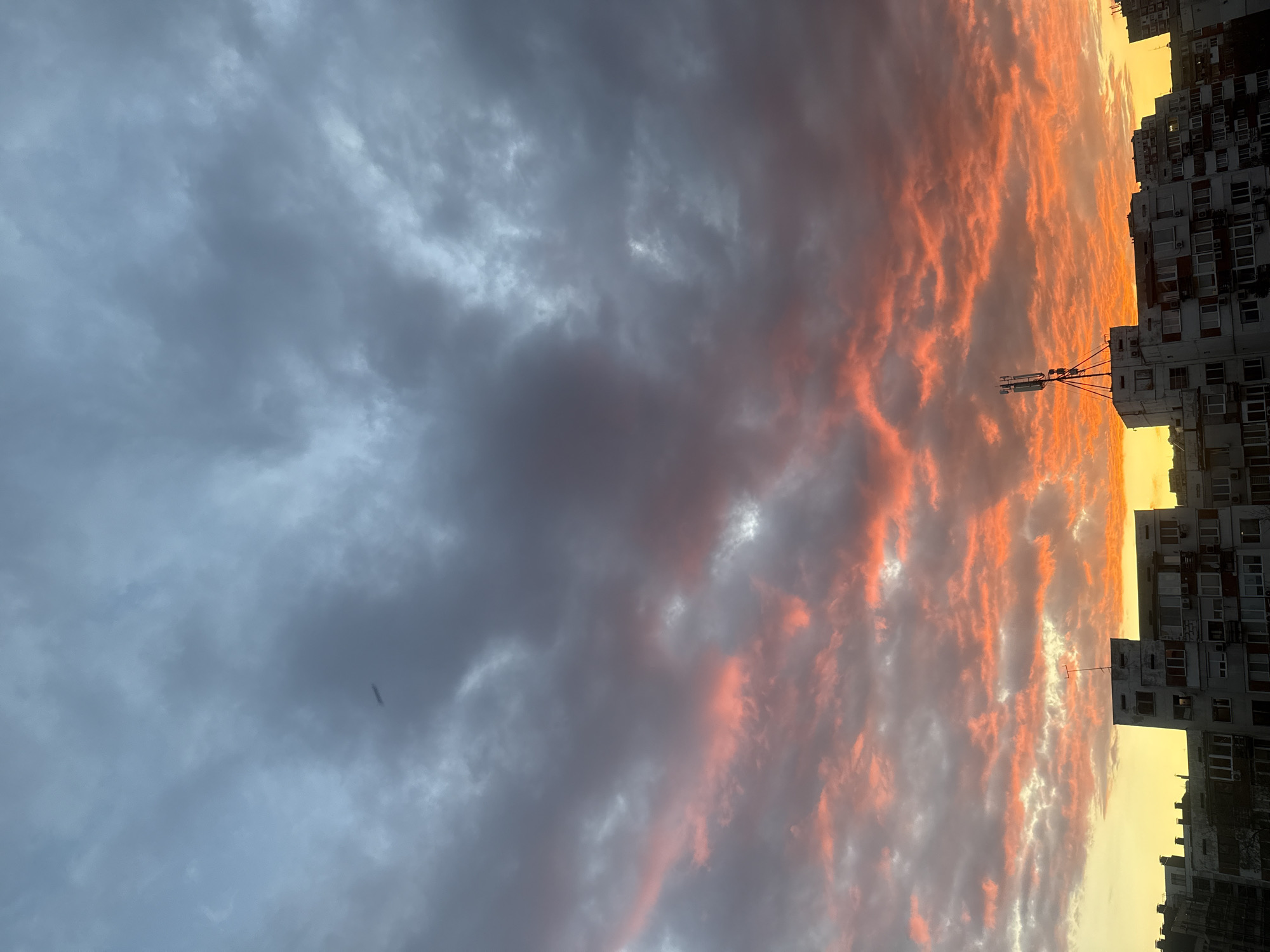 The image depicts a cloudy sky at sunset in what appears to be a city setting. To the left, the sky is filled with dense, grey clouds. To the right, the clouds are illuminated with orange and red hues from the setting sun. Buildings, likely residential apartments, are visible on the extreme right side of the image. A communication tower structure is silhouetted against the colorful sky. Based on the architecture and overall aesthetic, this could potentially be a residential district in the city of Sofia, Bulgaria. No people are visible in the image. The scene is static.