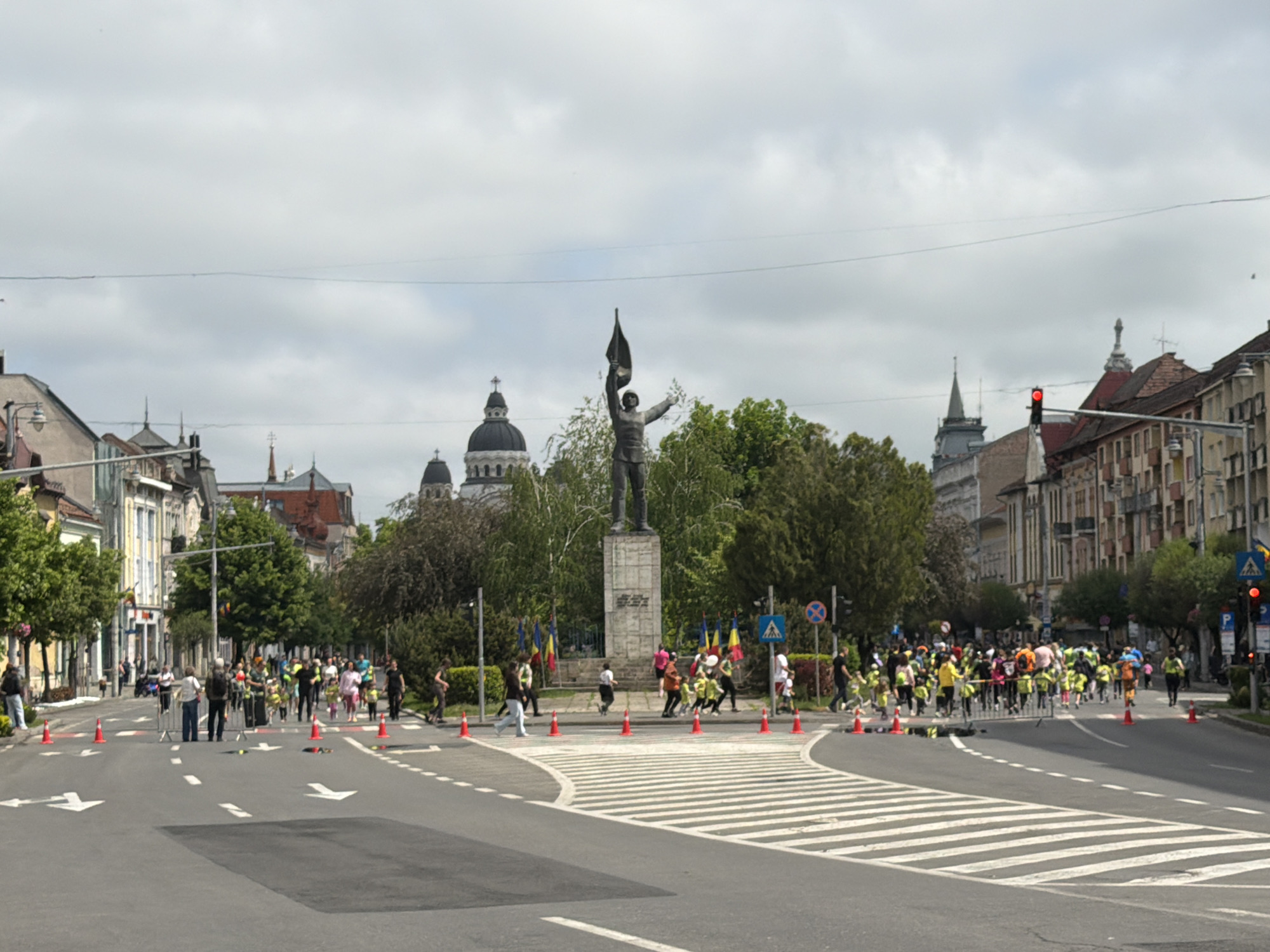 The image depicts a street scene in Târgu Mureș, Romania. A statue stands in a central median with Romanian flags around its base. Buildings line the street. A large group of people participates in an event that uses cones to mark pedestrian routes on the road. Pedestrians are visible on the sidewalk. A traffic light is on red. A crosswalk is present. The sky is overcast.