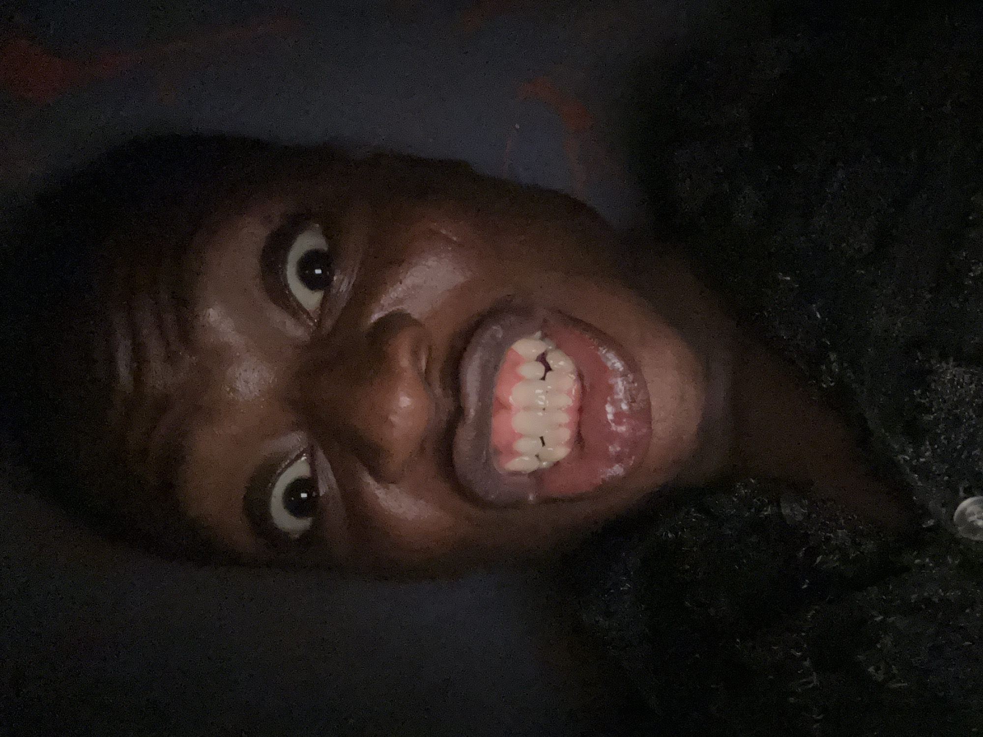 A close-up, low-angle shot depicts a person with dark skin, teeth bared in a grin, eyes wide and looking upwards. The background is dark and indistinct. Part of the person's clothing is visible on the right side of the image; it appears to be textured and dark. The specific location in the city of Unknown, Unknown is indeterminable based on the information provided.