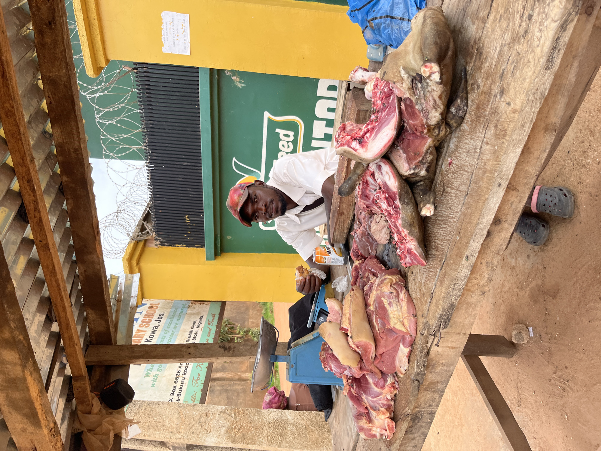 An outdoor meat stall in Jos, Nigeria, features a man in a white shirt and red cap behind a wooden table laden with raw meat. A blue scale is visible to the left, and a can of orange soda sits on the table. The stall is covered by a wooden and corrugated metal roof, and a yellow wall with green fencing is in the background. A sign for "ARY SCHOOL Kowa, Jos." is also visible. The ground is dirt, and a pair of black sandals are on the ground.