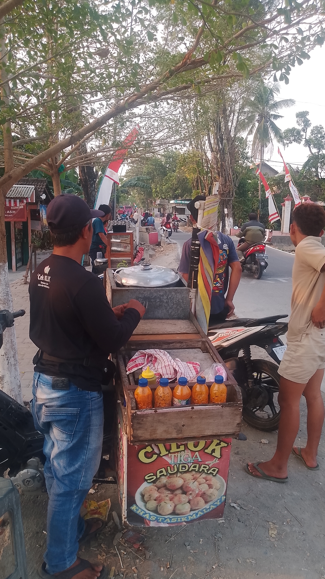 The image depicts a street scene in Karanglangu, Indonesia, likely during the daytime. In the foreground, a food vendor stands behind a cart selling "Cilok Tiga Saudara," a type of Indonesian street food. The cart is wooden and displays an image of the food item. Several bottles of sauce are arranged on top of the cart. A man wearing a black long-sleeved shirt and blue jeans is facing away from the camera, interacting with the cart. To the right, two young men are standing and observing. One is wearing a tan t-shirt and light-colored shorts, while the other is wearing a blue t-shirt and a colorful patterned cloth is tied around his head. In the background, a street is visible with several motorcycles and people, suggesting a busy area. The environment appears to be lined with trees and small shops. Indonesian flags are visible, adding a patriotic touch to the scene. The overall atmosphere suggests a casual, everyday moment in a local community.