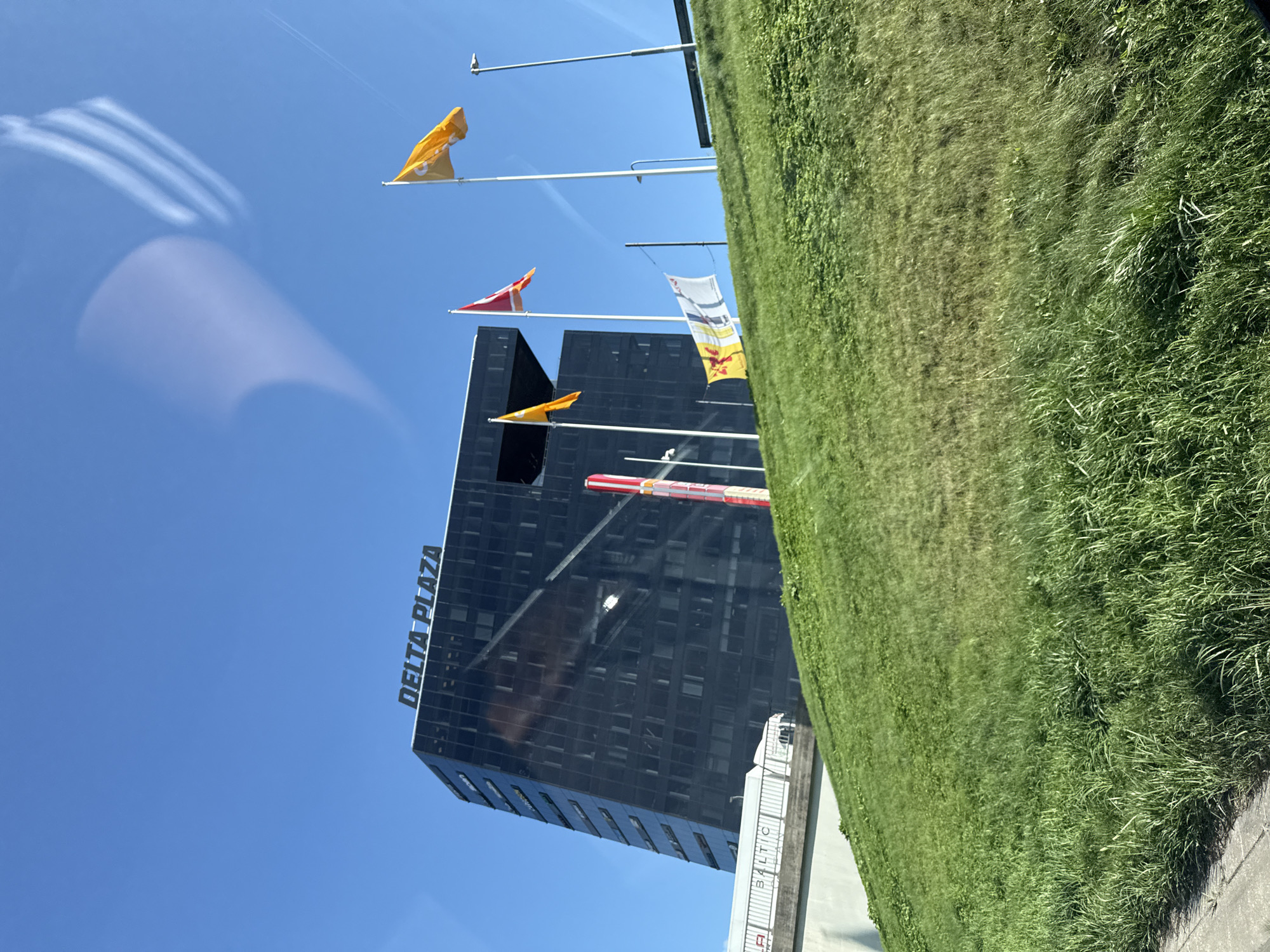 The image shows a modern, black glass-faced building with the text "DELTA PLAZA" visible on its top. The building is situated in Tallinn, Estonia. In front of the building, on a grassy incline, are several flagpoles with flags. Two prominent flags are yellow, and one is red. One yellow flag has a design that appears to be a coat of arms. To the left of the building, a white structure with the word "BALTIC" visible is present. The sky is clear and blue, with some faint cloud reflections on the building's glass facade, suggesting the photo was taken from a moving vehicle or through a window. The overall environment suggests an urban setting with some green spaces.