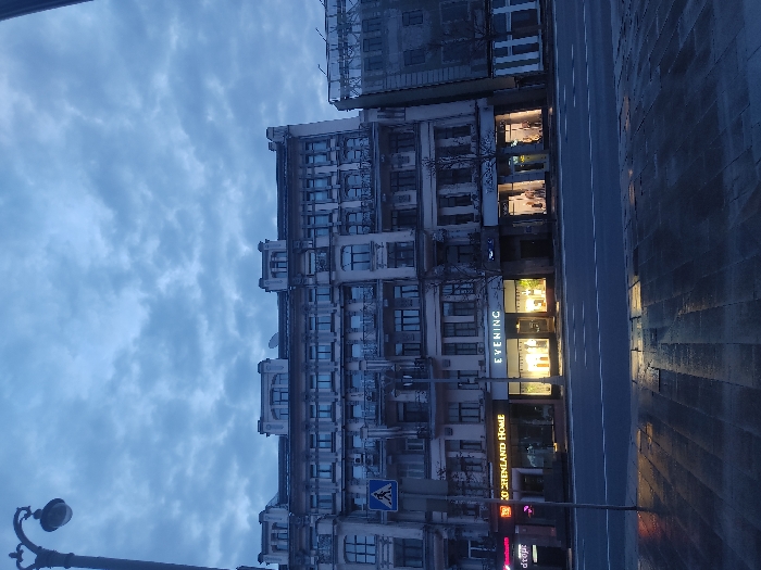 The image captures a street scene in Moscow, Russia. The street runs parallel to a multi-story building, which appears to be a mix of residential and commercial spaces. The building's facade is adorned with several balconies, windows, and architectural details.
Several storefronts are visible on the ground floor, with signs and lighting that suggest they are retail establishments. The lighting in these stores illuminates the adjacent sidewalk and street, creating a stark contrast with the overcast sky. A street sign with pedestrian crossing is present.
The road surface is dark and potentially wet, reflecting ambient light. A lamppost is also visible on the left side of the frame.
The overall atmosphere is muted, and the image suggests a time of day nearing dusk or dawn.