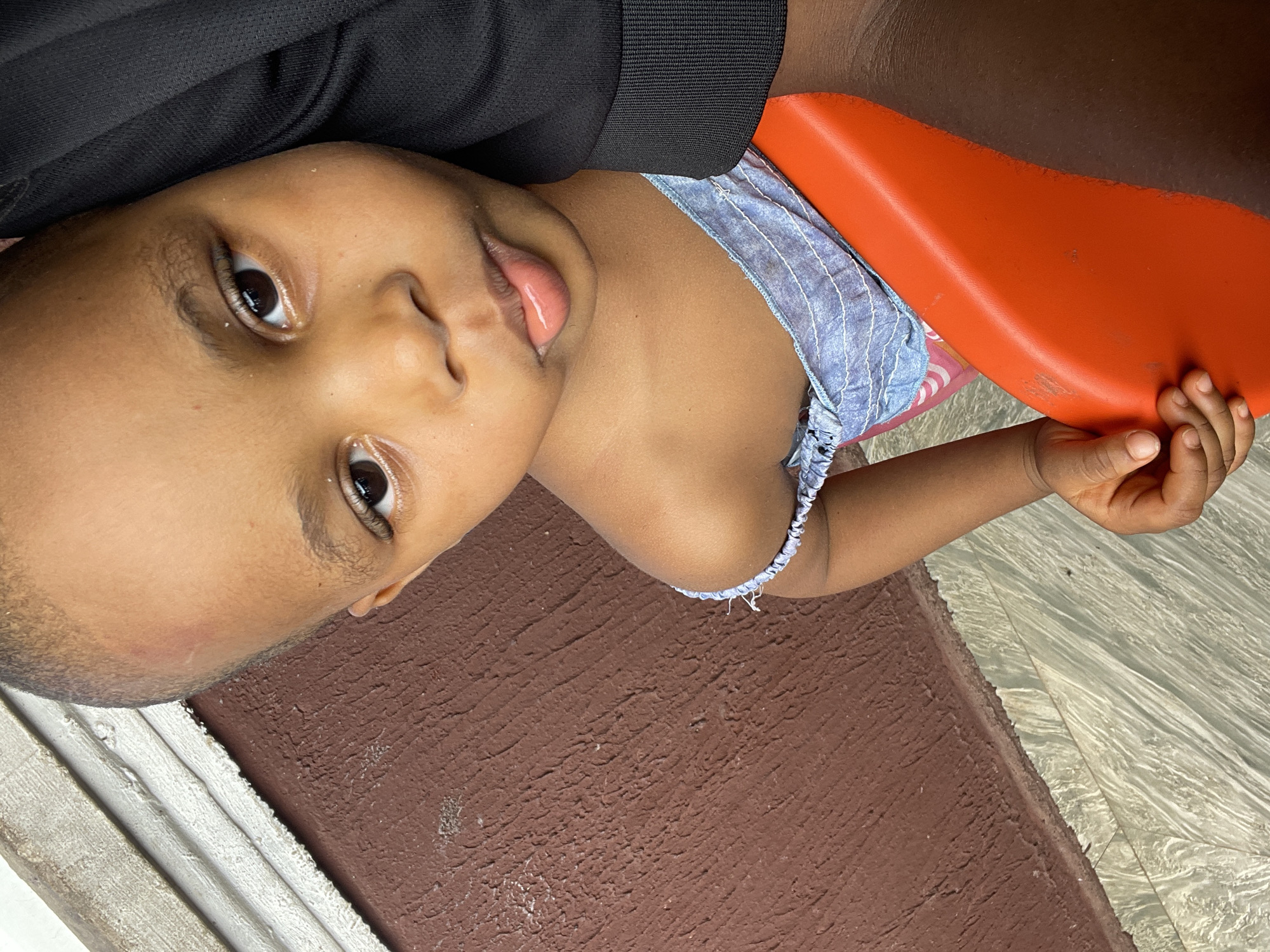 A child is lying on a textured surface that transitions from brown to a light, marbled pattern. The child wears a blue and white patterned top. Part of an orange object is visible near the child's body, held in the child's hand. The child is positioned next to a black cloth, which is situated alongside white molding. Based on the context, the location is assumed to be Onitsha, Nigeria.