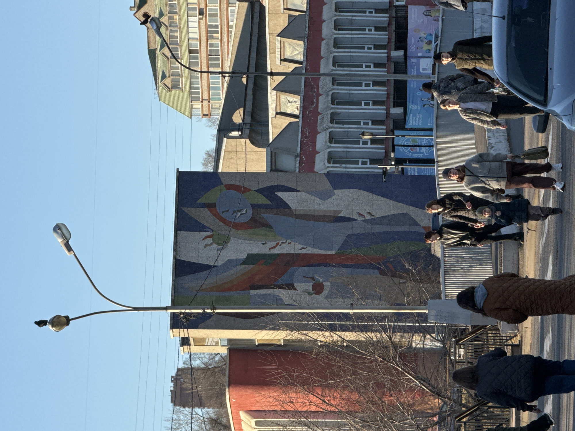 A street scene in Krasnoyarsk, Russia, features pedestrians crossing a zebra crossing in front of a large mosaic mural. The mural depicts abstract shapes, including a stylized sun, birds, and geometric forms in various shades of blue, red, and green. The mosaic covers a building facade. The buildings in the background include a taller residential structure with balconies and a partially visible, multi-story building with a green roof. Streetlights are visible to the left of the mural. Pedestrians include individuals wearing jackets, coats, and hats. A car is parked on the right side of the street, with its front portion visible. Bare branches of trees are seen in the lower portion of the image. The sky is clear and blue.