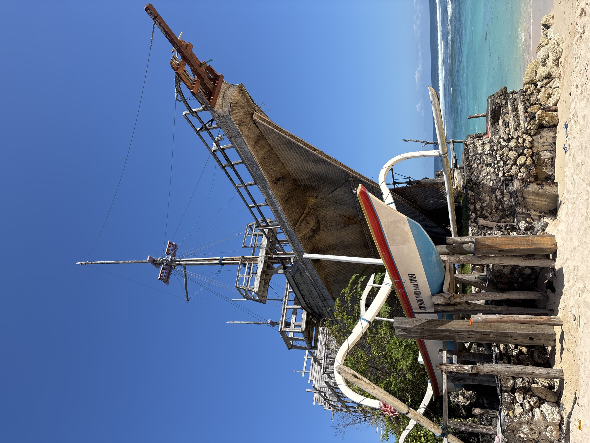 The image shows a traditional Indonesian outrigger boat, known as a "jukung," resting on a wooden structure on a beach. The jukung has a distinctive triangular sail made of woven material and tall outriggers. A second boat with a red and blue stripe rests next to it. The beach is sandy and adjacent to turquoise water. A rough wall of rocks and coral borders the beach. The sky is clear and blue. The location is likely in Kuta Selatan, Indonesia, known for its beaches and traditional boats.
