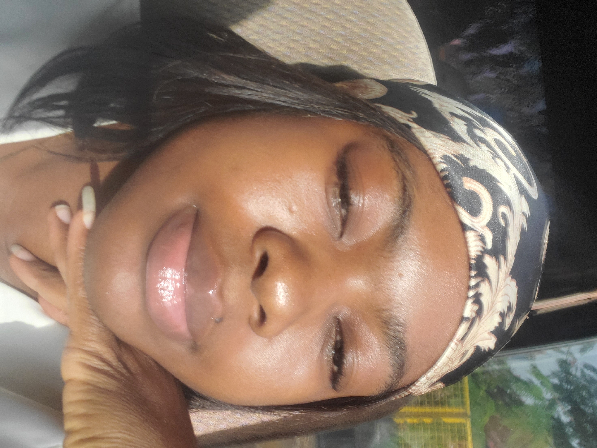 A close-up portrait shows a woman with dark skin and closed eyes. She is wearing a patterned scarf or headband. Her lips appear glossy. Her hand and a white cloth are visible near her neck. Behind her, a blurred scene suggests an outdoor setting with greenery and a yellow structure, possibly indicating the location is in the city of Gwagwada, Nigeria.