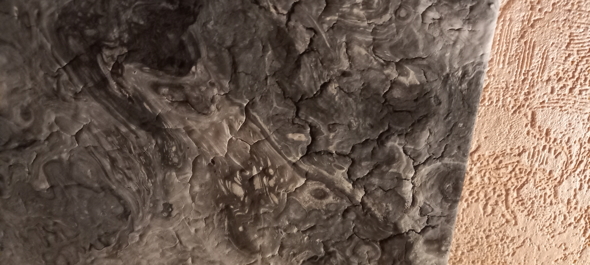 The image shows a close-up, abstract view of a textured surface. The upper portion features a dark gray, cracked, and swirled pattern resembling dried paint or a natural mineral formation. The texture is uneven, with raised ridges and deep fissures. Below this, a contrasting lighter, peachy-orange textured surface is visible, characterized by a consistent, bumpy pattern of raised ridges. This lower section appears more uniform in its texture compared to the upper part. There are no people, animals, or identifiable objects within the frame. The lighting suggests an indoor environment, possibly highlighting a wall or decorative element. The specific location in Kryvyi Rih, Ukraine, cannot be determined from the visual information provided in the image alone.
