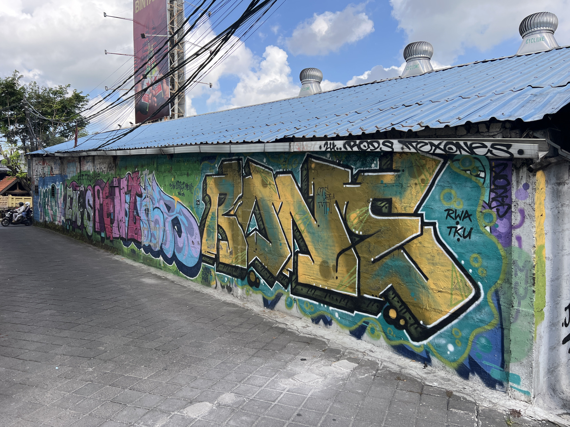 A street scene in North Kuta, Indonesia, features a gray-bricked street bordering a wall covered in graffiti. The graffiti is predominantly in green, yellow, pink, and blue. The yellow graffiti stands out prominently, with other various tags visible. Above the wall is a blue corrugated metal roof. On top of the roof are three metal cyclone vents. Power lines hang above the roof, and a tall billboard is visible in the background. A motorbike with two riders is parked in the distance on the left.