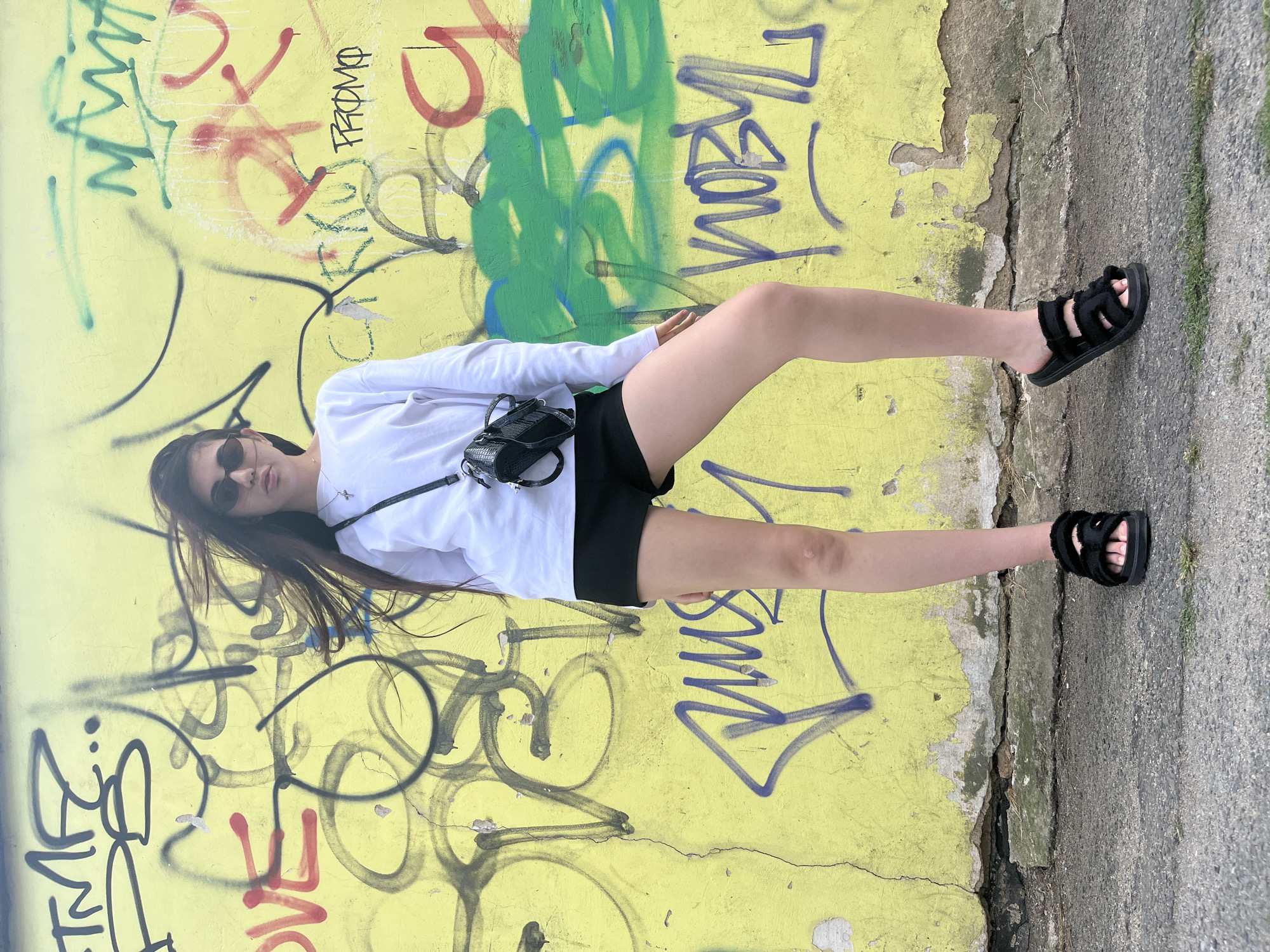 A young person with long brown hair and sunglasses stands facing forward in front of a yellow wall covered in graffiti. They are wearing a white long-sleeved shirt, black shorts, and black sandals. A small black purse with a textured design is slung across their chest. The graffiti on the wall includes various tags, scribbles, and some larger colorful markings. The ground in front of the wall is a rough, gray asphalt surface with some patches of green grass. The image appears to be taken outdoors in an urban environment.