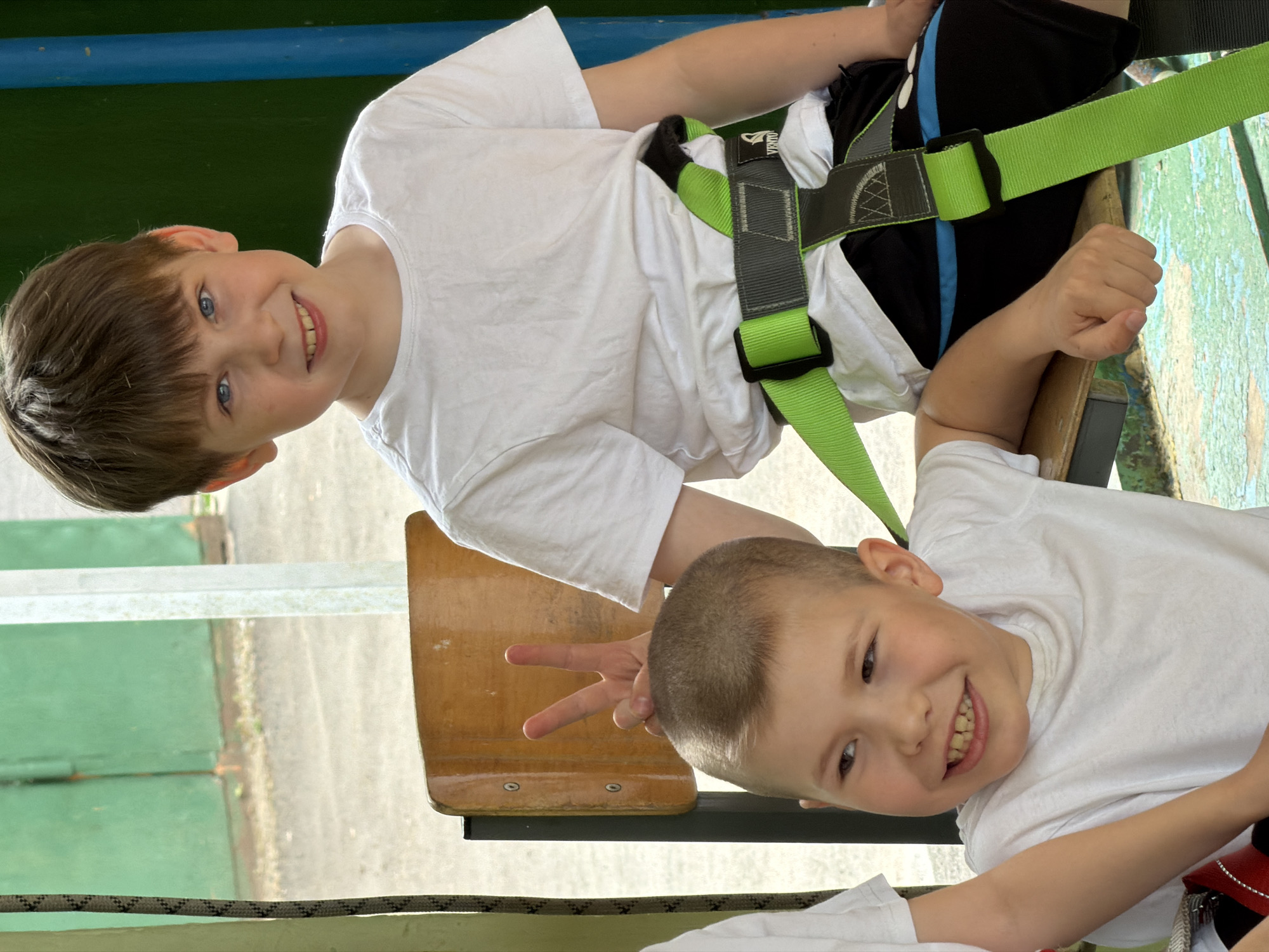 Two boys are posing for a photo while wearing safety harnesses. One boy is sitting on a wooden chair and making a peace sign, while the other boy is smiling at the camera. The location may be in Izhevsk, Russia, but there is no specific information to confirm this.