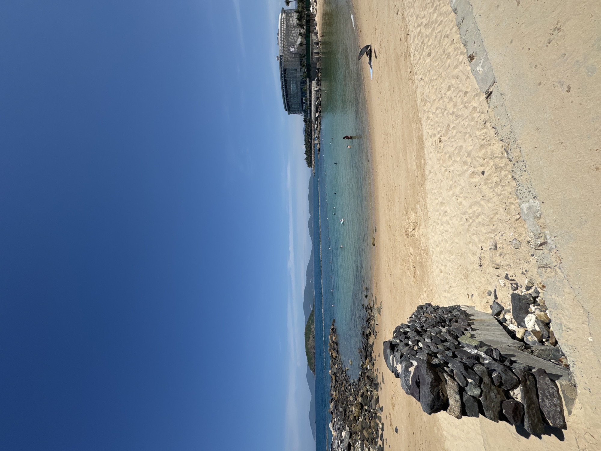 The image depicts a beach scene in Nha Trang, Vietnam, with a clear blue sky dominating the upper portion. In the foreground, a sandy beach stretches out, marked by a concrete path or seawall along the right side. A partially constructed or weathered stone structure sits on the left side of the beach. The water is a tranquil turquoise, with a rocky shoreline to the left and a small, tree-covered island in the middle ground. In the distance, a chain of hills or mountains forms the horizon. On the right side of the bay, a large, modern, multi-story building with a rounded facade and glass windows is visible, suggesting a resort or hotel. Several people are scattered throughout the water, some swimming and others wading. One person is visible in the shallow water on the right, near an open umbrella. The overall environment is sunny and idyllic, characteristic of a tropical beach destination.
