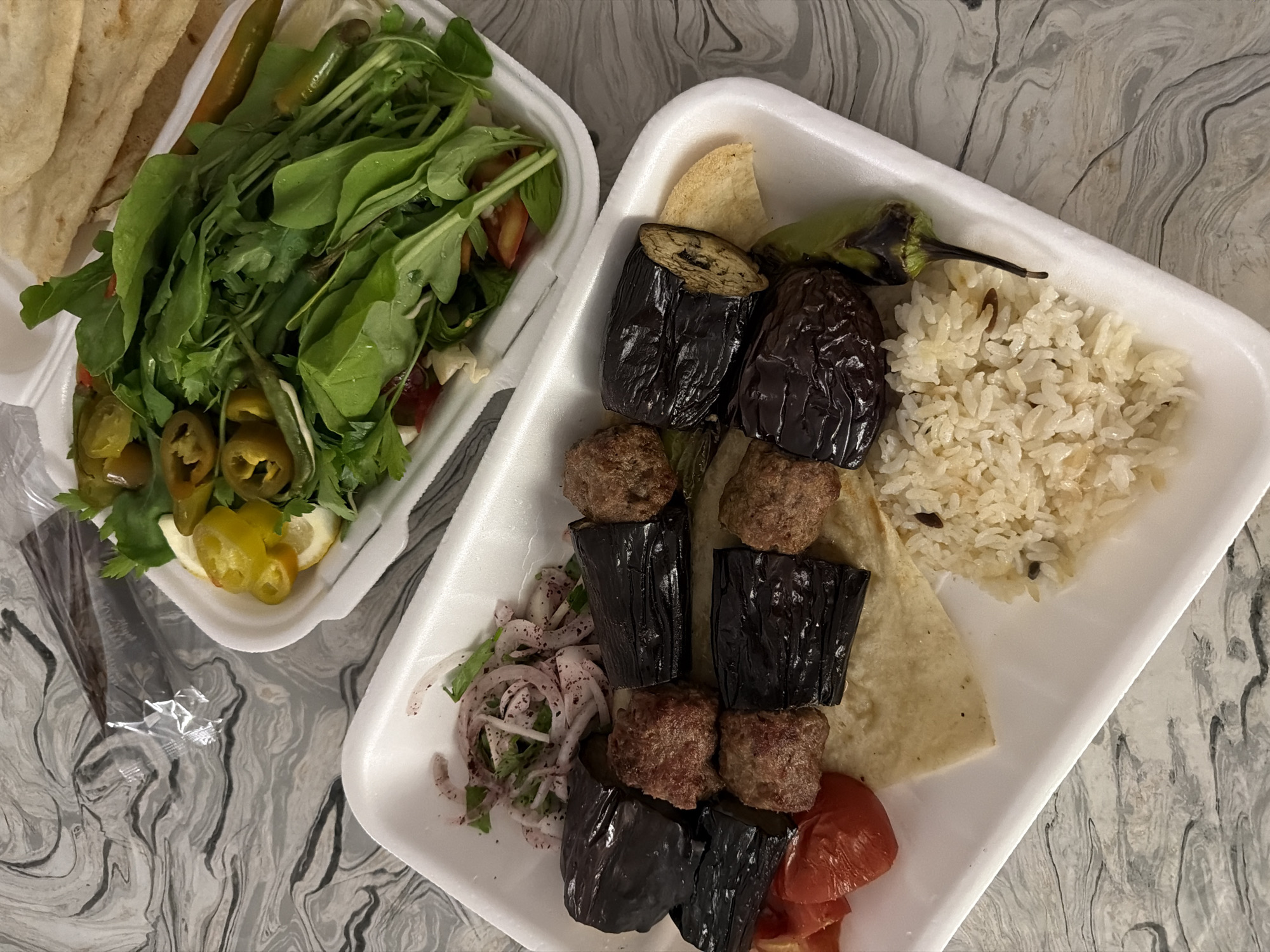 This image shows two white takeout containers filled with food, placed on a gray and white marbled surface. The container on the left holds a generous portion of green leafy salad, along with sliced jalapeños, lemon wedges, and what appear to be pickled peppers. Flatbread is stacked on the left side of this container.
The container on the right is filled with a meal that includes grilled eggplant pieces, meatballs, rice, a grilled green pepper, and sliced tomatoes. There is also a piece of flatbread under the meat and eggplant. Thinly sliced red onions with herbs are visible to the left of the grilled items.
The food appears to be a takeaway meal, likely from a restaurant. The lighting suggests it could be daytime. There are no people or animals visible in the image. The overall scene depicts a meal ready to be eaten.