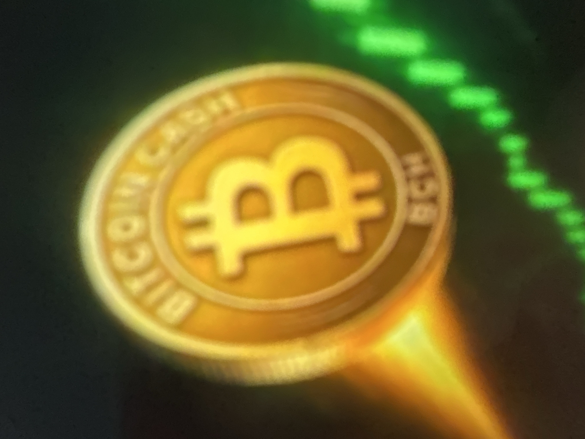 The image displays a close-up, slightly blurred view of a Bitcoin Cash (BCH) coin. The coin is golden and features a prominent "B" symbol in its center. Text surrounding the "B" is partially visible, with "BITCOIN CASH" appearing on the upper curve and "BCH" on the lower curve. To the left of the coin, a bright, glowing orange and yellow light emanates, suggesting a rising value or positive momentum. To the right and below the coin, a green line graph with rising bars is visible, further symbolizing financial growth. The background is dark and gradually transitions from black to a dark green hue, creating a dramatic contrast that emphasizes the coin and the financial indicators. The image does not contain any elements that would indicate a location in Gelendzhik, Russia, or any specific urban environment.