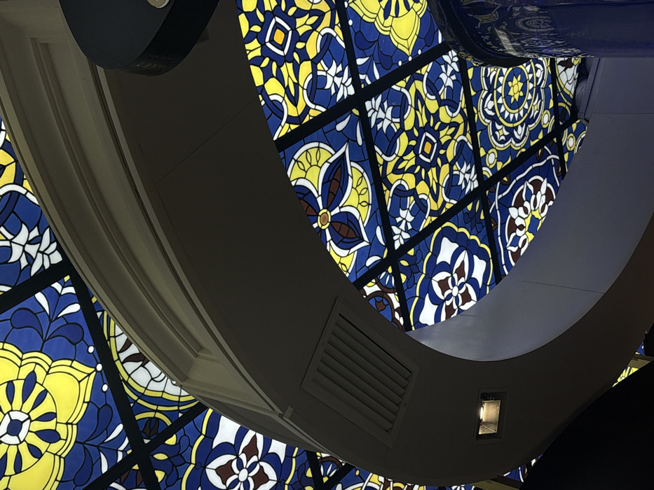 Looking upwards, a view is framed by a curved, grey-toned architectural structure featuring ventilation grilles and an inset light. Beyond the structure, a colorful patterned ceiling is visible, comprised of blue, yellow, and white floral designs separated by dark lines. The perspective is from below, highlighting the ceiling’s intricate details.