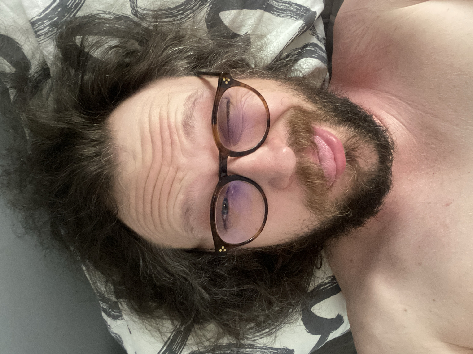 In Rusanovo, Russia, a person is lying on a bed with their head on a pillow. The person has long, dark, messy hair and is wearing eyeglasses with tortoiseshell frames. Their face is partially obscured by their hair, and they are winking with one eye while sticking out their tongue. The person has a beard and appears to be shirtless. The pillow has a black and white patterned design. The lighting suggests an indoor setting.