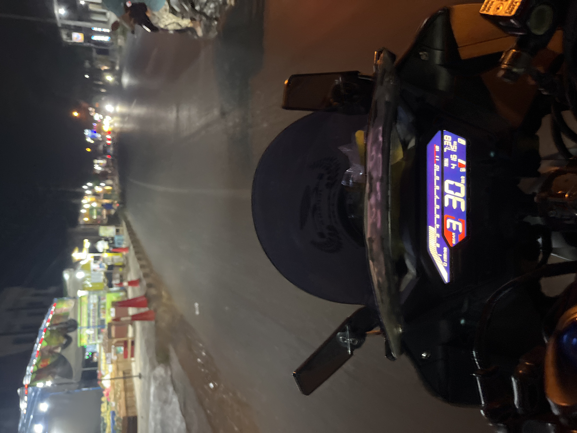 The image shows a first-person perspective from a motorcycle at night, likely in Pecangaan Wetan, Indonesia, given the context. The rider's digital display shows a speed of 30 km/h and the gear is set to "3". The time is displayed as 7:38. In the foreground, a dark windshield is visible, along with a black motorcycle dashboard. The road ahead is illuminated by streetlights and the headlights of other vehicles. On the left side of the road, there are several food stalls or shops with bright lighting, displaying goods for sale. Red traffic cones are placed along the edge of the road. The overall environment suggests a bustling street scene at night.