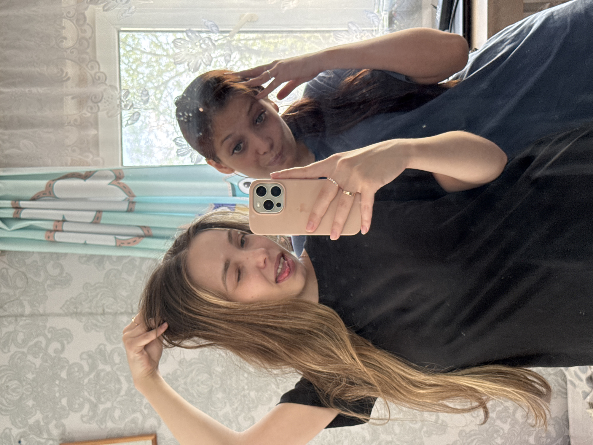 A photograph captures two women reflected in a mirror. The primary subject, in the foreground, holds a phone with a beige case, obscuring the camera's details. She is fair-skinned, has long, highlighted brown hair, and wears a black shirt. Her face is animated with a wink and a protruding tongue. Behind her, the reflection reveals another woman with darker skin and reddish-brown hair, wearing a blue shirt. She is holding her hair with one hand and looking towards the camera. The background features a patterned wallpaper and a partially visible window with sheer curtains, suggesting an indoor environment. The light suggests that it's daytime. Кемуль, Russia, is not directly discernible within the visual context of the image.