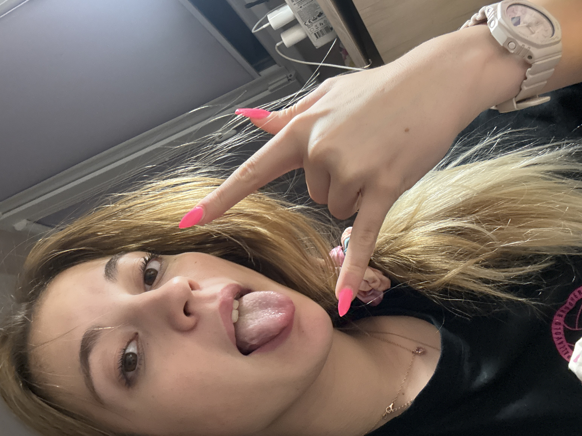 A young woman is posing for a selfie, sticking her tongue out. She has blonde hair and is wearing a black shirt. Her fingernails are painted bright pink. She is making a hand gesture. She is wearing a watch on her left wrist and a gold necklace. The background includes a window frame, wall plugs and a dark surface. There is no indication of the location being in Sofia, Bulgaria.