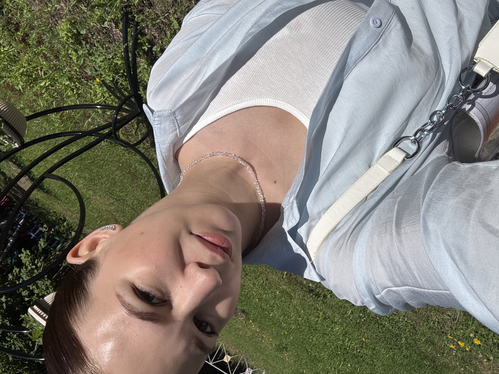 The image is a selfie taken outdoors on a sunny day. The subject is a young woman with her hair pulled back, wearing a light blue open button-up shirt over a white ribbed tank top. She has a delicate necklace with small beads around her neck and is wearing a stud earring in her visible ear. She has a white purse with a chain strap slung over her shoulder.
The background consists of green grass with scattered small yellow flowers and the dark metal frame of what appears to be outdoor furniture. The lighting suggests it is daytime, with sunlight casting shadows. The overall impression is that of a casual outdoor setting, possibly a park or a backyard in Chaikovsky, Russia, given the request.