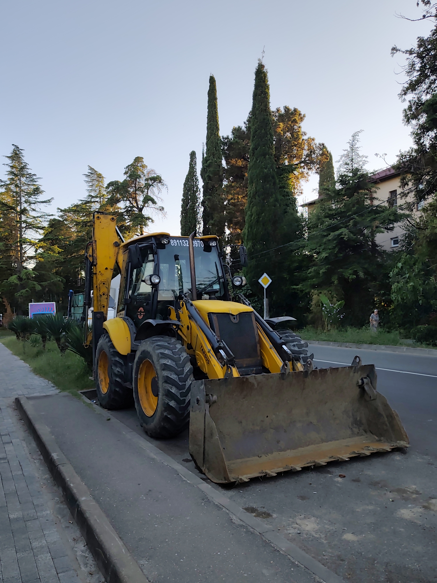 An outdoor, eye-level shot captures a yellow backhoe loader parked on the side of a road in Sochi, Russia. The construction vehicle is positioned on a paved area next to a sidewalk made of gray interlocking pavers. The backhoe's large, dirt-covered bucket is extended forward and rests on the asphalt. The vehicle has prominent black tires with yellow rims.
Behind the backhoe, a paved road stretches into the distance. The environment is lush with greenery; tall, slender cypress trees dominate the landscape, interspersed with broader, leafy trees. Some of these trees are illuminated by the warm glow of what appears to be late afternoon or early evening sun. A multi-story building with a light-colored facade and dark windows is visible behind the trees on the right side of the image. A yellow diamond-shaped road sign with a black border is positioned near the backhoe. A single person, wearing a patterned dress, is visible in the distance, standing near the edge of the trees on the right side of the road. A partially visible sign with colorful graphics is located to the left of the backhoe. The sky is clear and a pale blue.