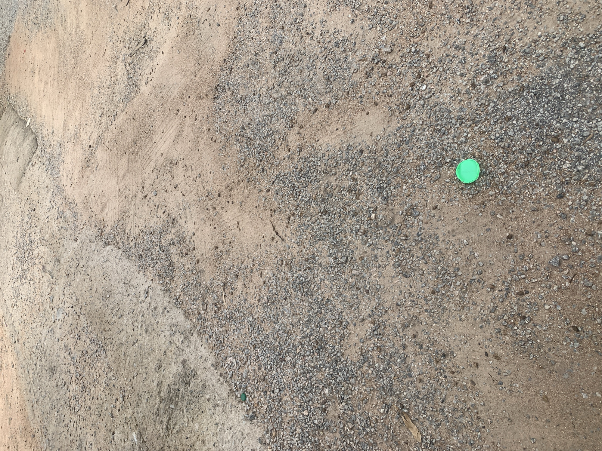 The image shows a top-down view of a dirt and gravel surface, likely a path or unpaved road. The ground is composed of small, greyish pebbles mixed with fine sand. In the upper left quadrant, there is a patch of smoother, lighter-colored dirt, possibly indicating a more compacted area or a different type of soil. The texture of the ground is rough and uneven.
A single, bright green plastic bottle cap is visible in the right-center of the image, resting on the gravel. Its color stands out against the muted tones of the ground. Scattered across the surface are a few small twigs and dried leaves, suggesting an outdoor environment. The lighting appears to be natural daylight, casting no distinct shadows, which could indicate an overcast sky or midday sun.
There are no people or animals present. The scene is one of stillness and a lack of human activity, other than the presence of the discarded bottle cap. The location cues, such as the composition of the ground, are consistent with a rural or undeveloped area. No text is visible in the image. The overall impression is of a simple, utilitarian outdoor space.