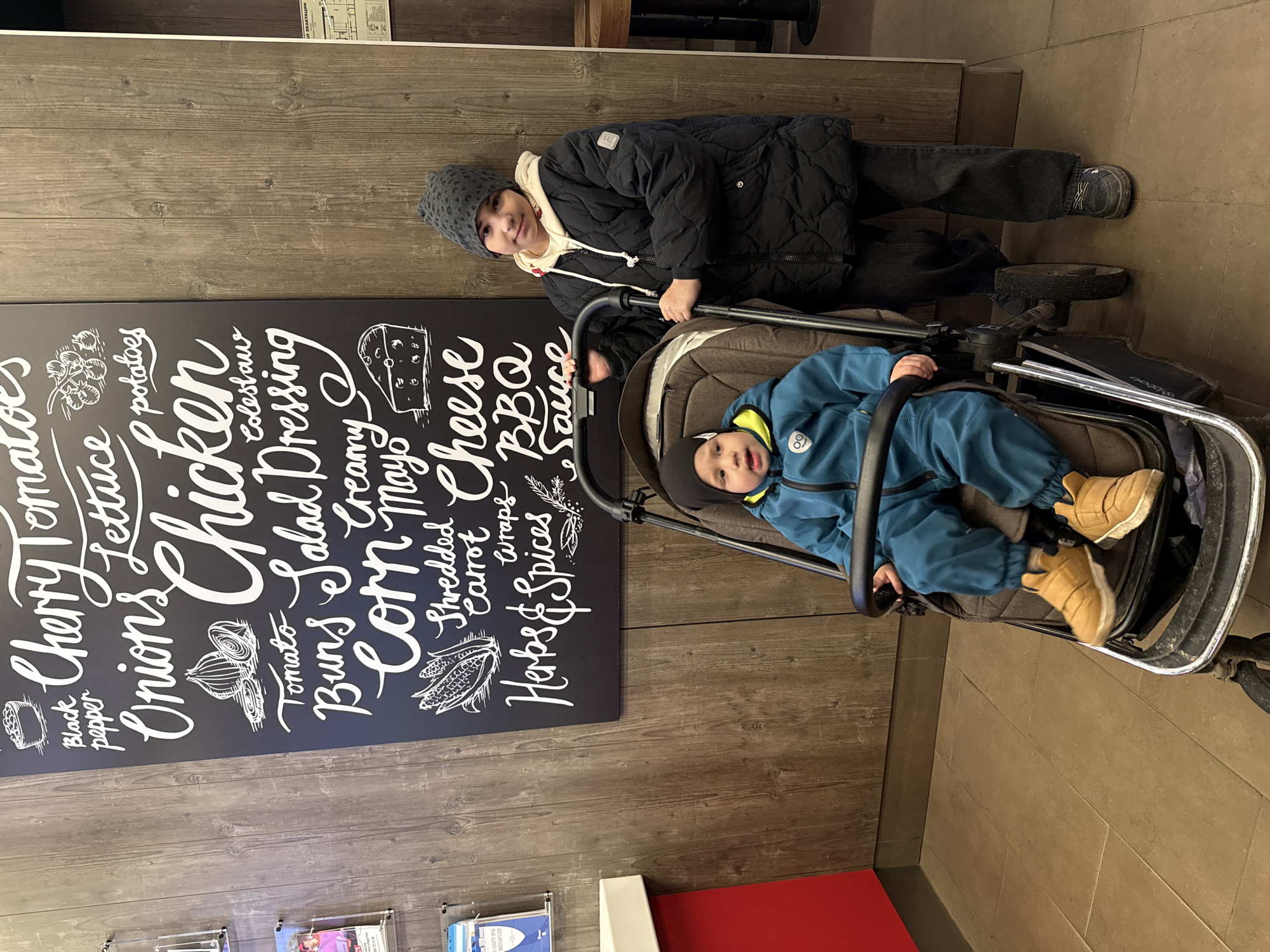An indoor portrait-oriented image captures two children in what appears to be a cafe or casual dining establishment. An older child, dressed in a dark padded winter jacket, light hoodie, dark trousers, and a grey knitted beanie, stands on the left, pushing a stroller. This child looks directly at the camera with a gentle smile.
Seated in the stroller is a younger child, wearing a blue padded winter suit, a dark beanie, and light brown boots. This child also looks towards the camera with an attentive expression. The stroller is dark with a robust frame and wheels.
The background features a wooden-paneled wall, a dark chalkboard-style sign with white, decorative text, and a light-colored tiled floor. The chalkboard sign, resembling a menu, lists various food ingredients and dishes including "Black pepper," "Cherry Tomatoes," "Onions," "Lettuce," "potatoes," "Chicken," "Tomato," "Buns," "Salad Dressing," "Creamy," "Corn," "Mayo," "Shredded Carrot," "Cheese," "Wraps," "BBQ," "Herbs & Spices," and "Sauce." It also features illustrations of onions, corn, and a cheese block.
In the upper right corner, a partially visible poster displays Cyrillic text, which reads "ОБЪЕДИНЕНИЕ ПОТРЕБИТЕЛЕЙ" (Consumers' Association). Several brochures in clear holders are visible on the lower left wall, and a red surface is partially seen on the floor. The children's warm clothing suggests cold weather outside, consistent with an indoor setting in Rusanovo, Russia.