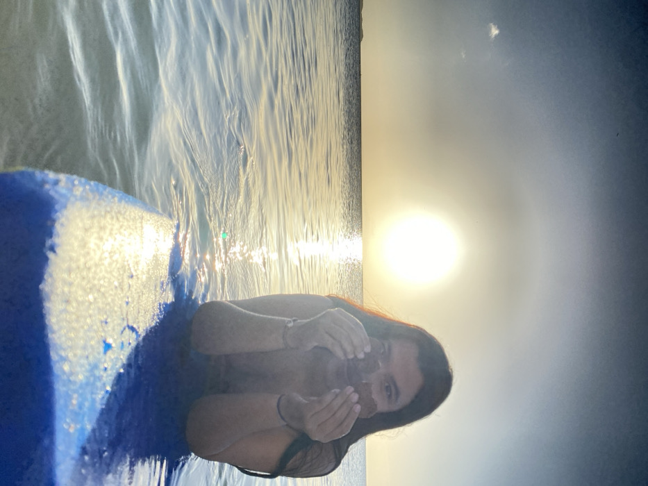 A woman with long dark hair and glasses is in the water. She holds something small in her hands near her face. She's on a blue object, possibly a float or raft. The sun is behind her, creating a bright halo effect and light reflecting on the water's surface. The sky is partly cloudy, transitioning from light to darker blue. The water appears calm and shimmering.