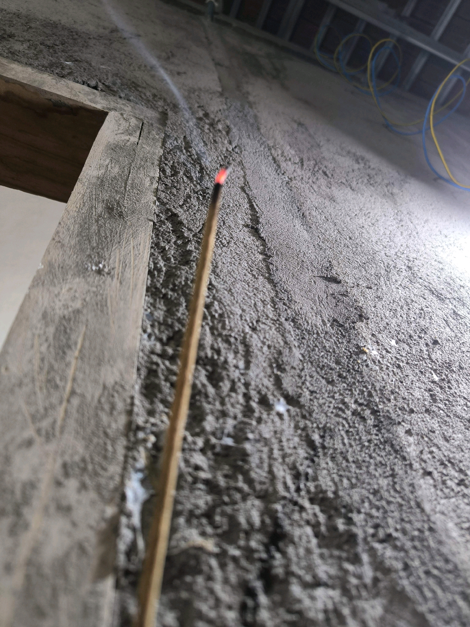 An incense stick is burning and smoking against a textured concrete wall. The incense stick is made of wood and is positioned vertically, with its glowing red tip and wisps of smoke clearly visible. To the left of the incense stick is a rough, wooden beam, possibly part of a doorway or construction frame. The wall itself appears to be in a state of construction or disrepair, with a rough, uneven surface. In the background, above the wall, there are visible electrical wires, coiled and colored yellow and blue, suspended from a metal framework, suggesting an unfinished building or infrastructure in Tabanan, Indonesia. The lighting is dim, casting shadows and highlighting the textures of the wall and the smoke.
