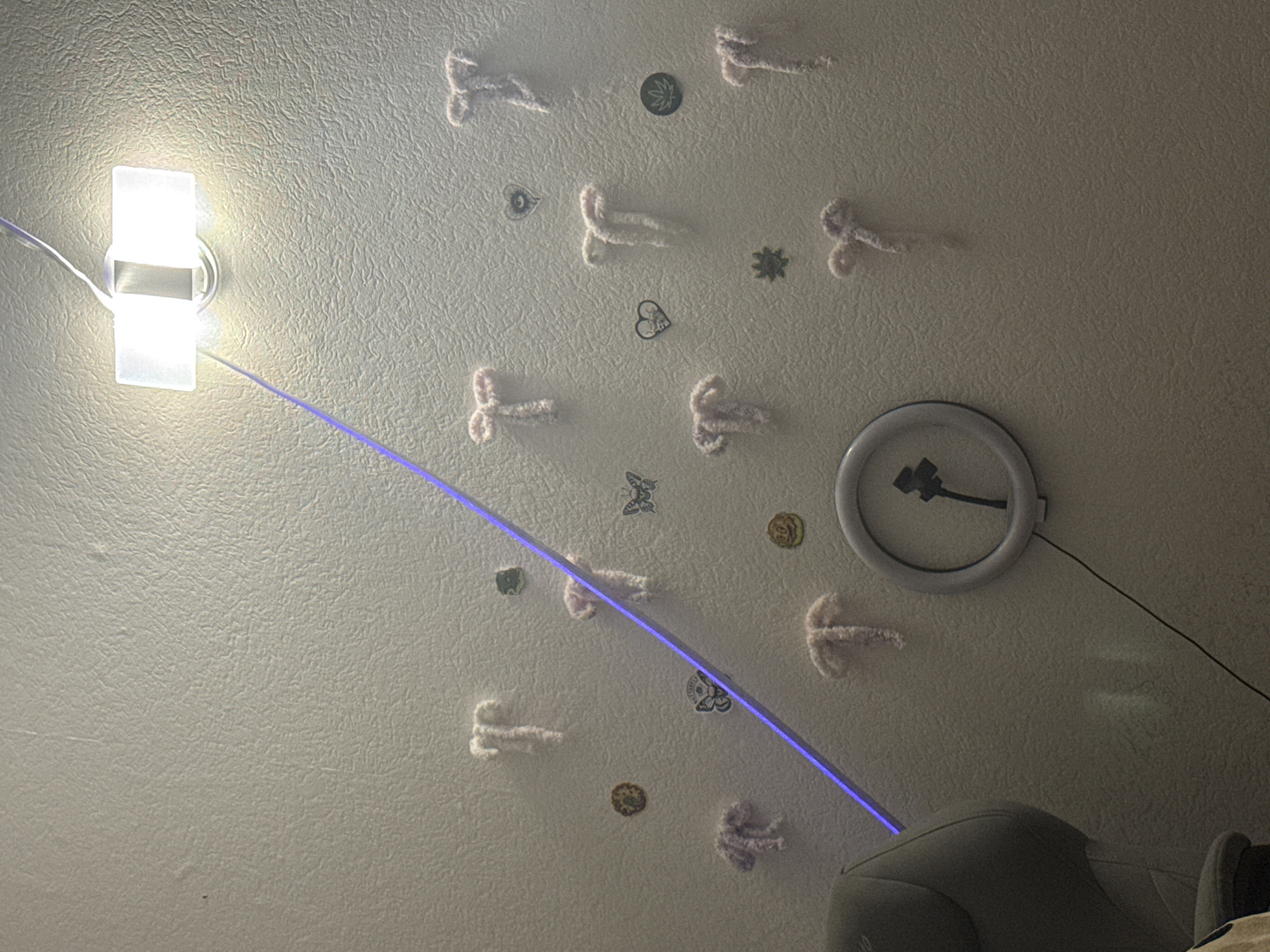 This is a medium shot, eye-level, indoor photograph of a textured white wall in an unknown location. The wall is adorned with several pink fuzzy pipe cleaner figures, each resembling a stylized uterus or butterfly. Scattered among the figures are small stickers of a marijuana leaf, a butterfly, a heart with an eye, and a circular brown design. A bright overhead light fixture illuminates the top right portion of the wall. A thin, bright blue laser beam cuts diagonally across the scene from the top left to the bottom left. To the left of the frame, a portion of a grey office chair is visible. In the lower center of the image, a circular ring light, likely a selfie light, is attached to the wall. A black cord extends from the ring light downwards and to the left. The overall impression is of a decorated wall, possibly in a bedroom or personal space, with a decorative laser light feature.