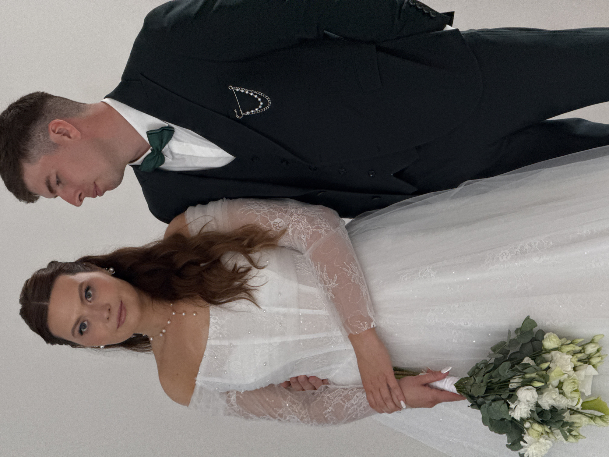 This portrait captures a newlywed couple in what appears to be a studio setting, likely in Chaikovsky, Russia. The image is oriented vertically, with the subjects appearing rotated 90 degrees counter-clockwise.
The man, presumably the groom, stands on the left, looking down with a contemplative expression. He has short, dark hair and is dressed in a dark suit (possibly dark green or black), a crisp white shirt, and a dark green bow tie. A decorative silver-toned lapel pin with pearls and chains adorns his left lapel.
The woman, the bride, is positioned slightly in front and to the right of the man, looking directly at the viewer with a calm and gentle expression. She has long, wavy dark brown hair, styled partly pulled back. She wears an elegant white wedding gown featuring off-the-shoulder styling and long, sheer lace sleeves. The bodice of her dress is embellished with delicate pearls. Her accessories include pearl drop earrings and a pearl necklace. Her nails are manicured with white polish. She holds a beautiful bouquet of white and light green flowers, including what appear to be eustoma/lisianthus and eucalyptus leaves, wrapped in white ribbon.
The background is a plain, light-colored surface, suggesting an indoor professional photoshoot environment. No specific time of day or weather conditions are discernible. There is no visible text in the image. The overall impression is one of a formal and intimate wedding portrait session.
