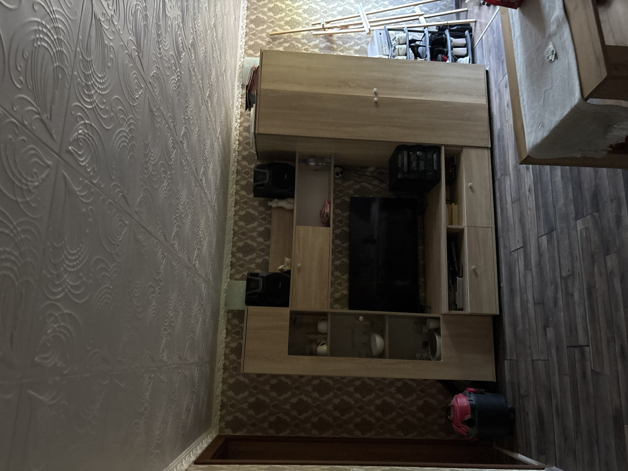 The image shows an interior view of a room. The walls are covered in a patterned wallpaper with a floral motif in shades of brown. The ceiling is textured with a white, embossed pattern resembling swirling designs.
In the center of the room, there is a large wooden entertainment unit with multiple shelves and cabinets. A black flat-screen television is set into the unit. Above the television, there are shelves displaying various items including cups, a bowl, and two black speakers. To the right of the television, there is a black electronic device with multiple buttons and a display.
To the left of the entertainment unit, a red and black shop vac is positioned on the floor.
To the right of the entertainment unit, there is a tall, light-colored wooden wardrobe with two doors. Above the wardrobe, a stack of books or papers is visible.
Further to the right, a wooden easel stands with a canvas on it, and to the right of the easel, a multi-tiered shoe rack is filled with shoes.
In the foreground, a wooden table with a light-colored tablecloth is partially visible. The floor is covered with dark wooden laminate flooring.
The scene depicts a living area, possibly in a home, in Redina, Bulgaria. There are no people visible in the image. The lighting suggests it is daytime, with natural light entering from an unseen source.