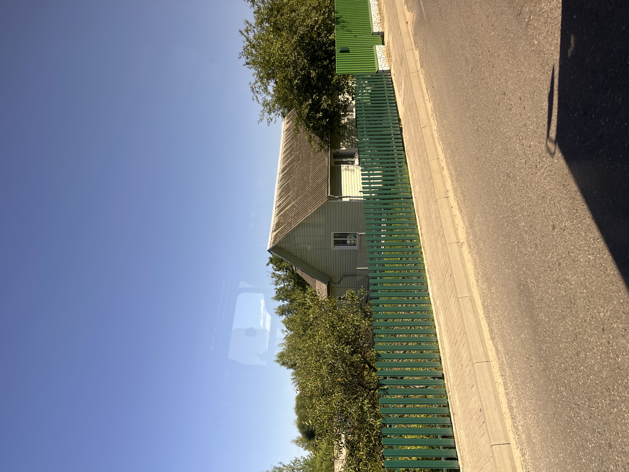 The image shows a residential street in Pastavy, Belarus, on a clear day. A light green house with a grey, corrugated roof stands behind a tall, bright green metal fence. The fence runs along a paved sidewalk, which is adjacent to a paved road. Trees and foliage are visible behind the fence, and the sky is a clear, bright blue. The overall scene depicts a quiet, suburban environment.