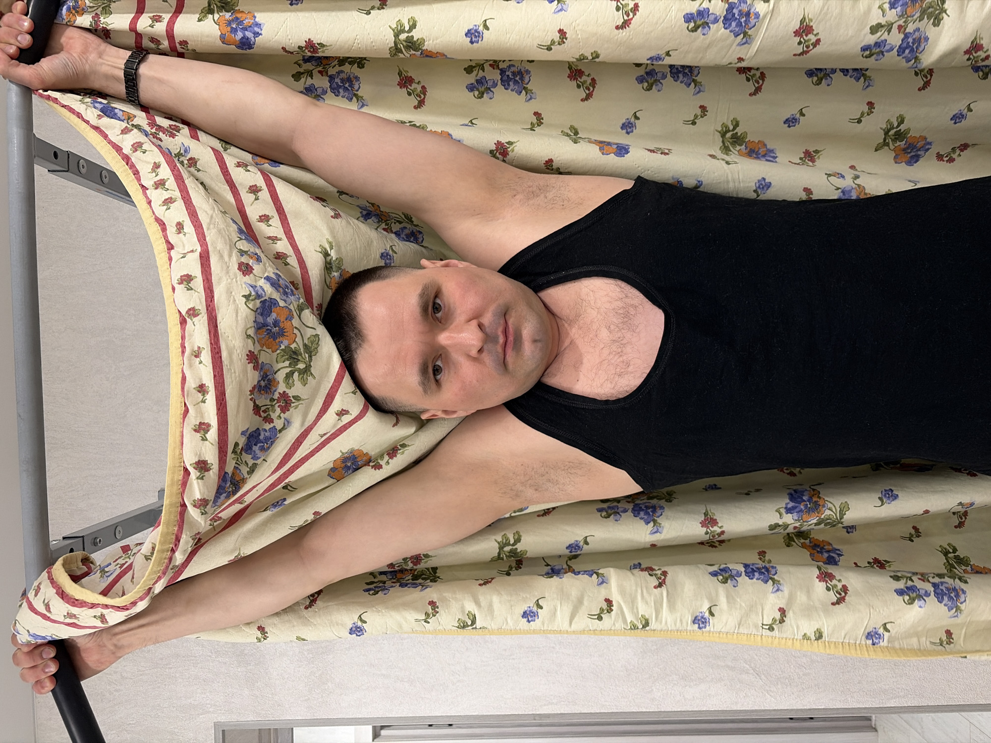 A man lies on a floral patterned bed, wearing a black tank top. He has short hair and his arms are extended above his head, holding onto the bed frame. He is looking directly at the camera. The location is potentially in Sarapul, Russia.