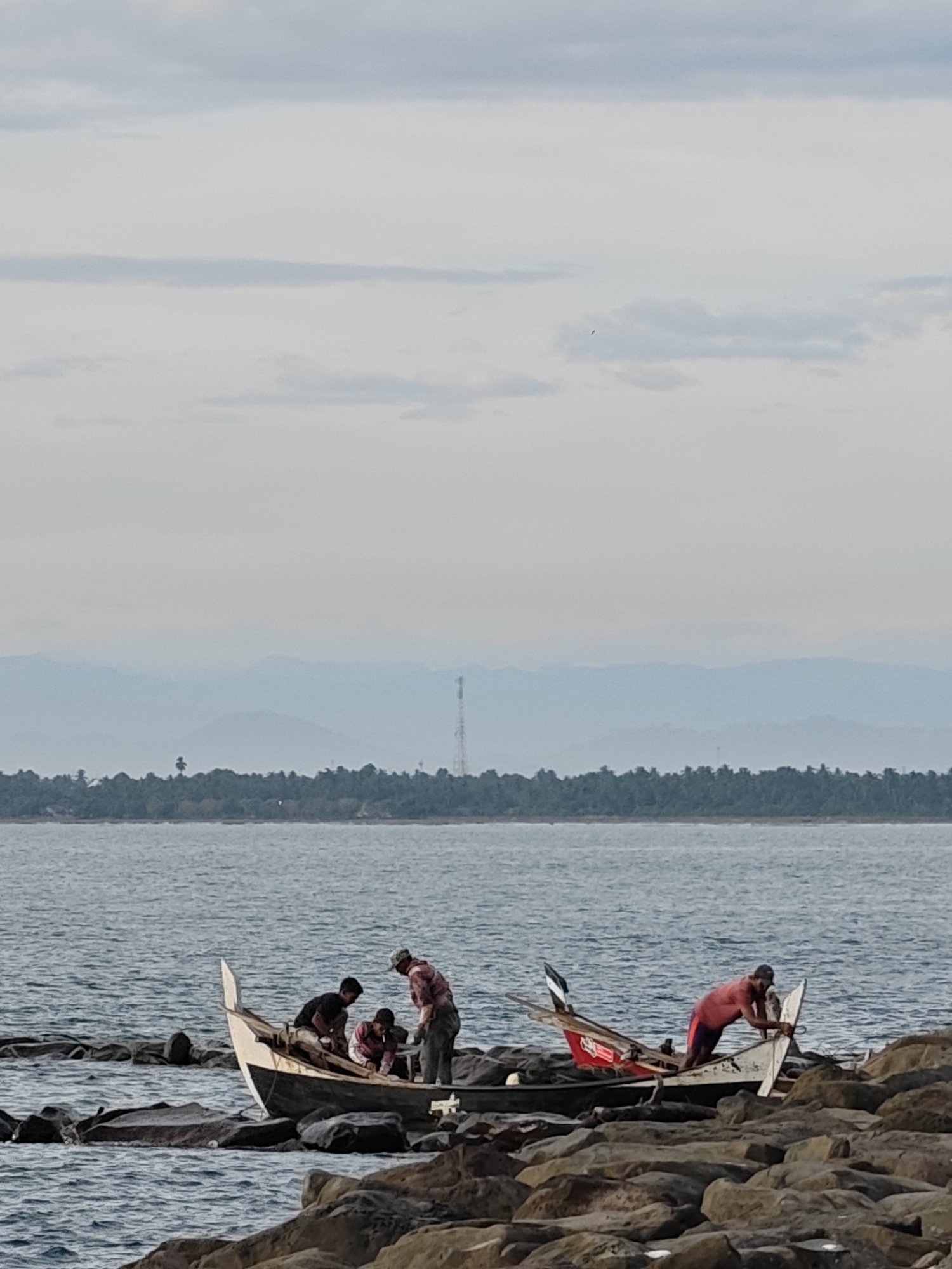 On a rocky shoreline, likely in the city of Lhokseumawe, Indonesia, two boats are situated near the water's edge. Four people are congregated around the first boat, while one person stands near the second. The sea extends towards a distant coastline with mountains visible in the background under a cloudy sky. A tall tower is also visible on the coastline.