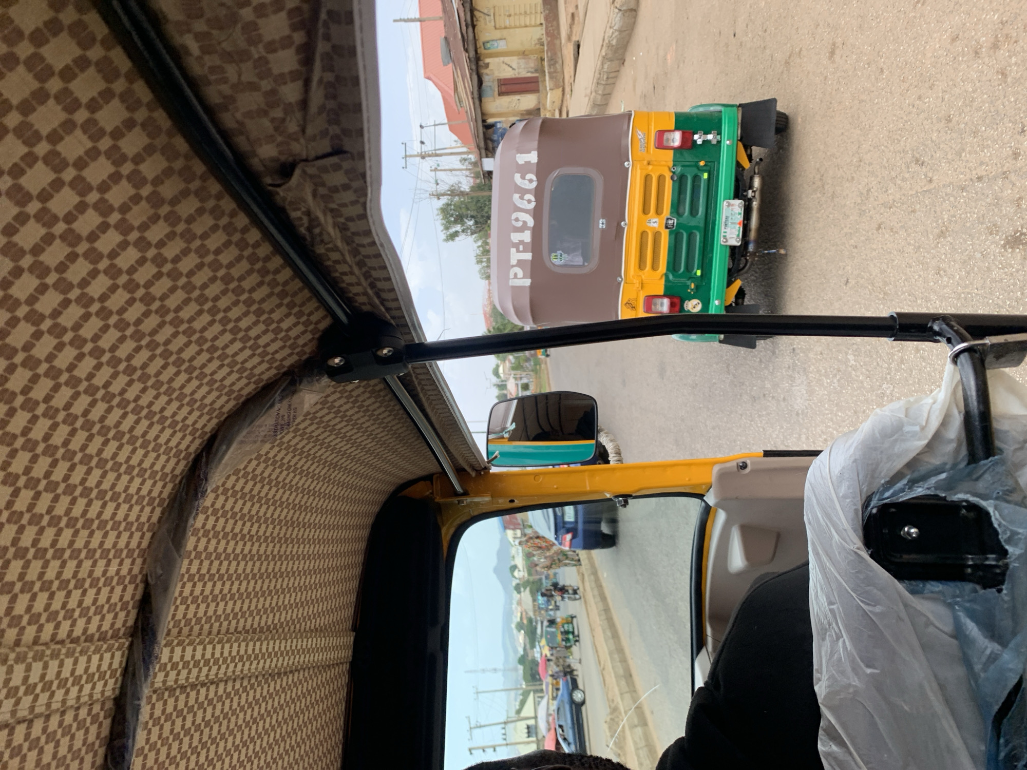 The image shows a view from inside a vehicle, possibly a tricycle or "Keke Napep", in Jos, Nigeria. The patterned interior of the vehicle's roof is visible on the left. Through the front window, another similar vehicle is seen with a license plate "PT-19661". The streetscape outside the window includes other vehicles and buildings. A side mirror reflects a busy street scene in Jos.
