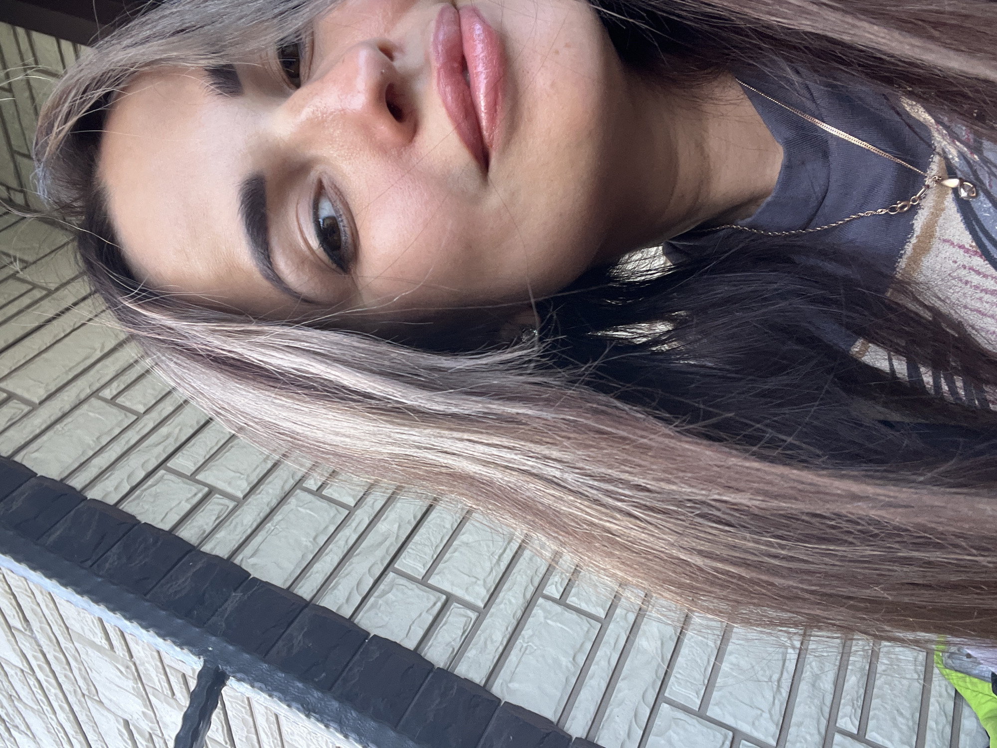 A close-up, low-angle selfie shows the upper half of a young woman's face and her hair, which is styled with blonde highlights. She is wearing a dark t-shirt and a delicate gold necklace. Her lips are glossed a rosy pink, and her eyebrows are defined and dark. Her right eye is looking directly at the camera, while her left eye is partially obscured by her hair. The background is a textured, light-colored brick wall. The lighting suggests it may be daytime, possibly outdoors, given the ambient light and the textured wall which could be part of a building's exterior. There is no visible text in the image.