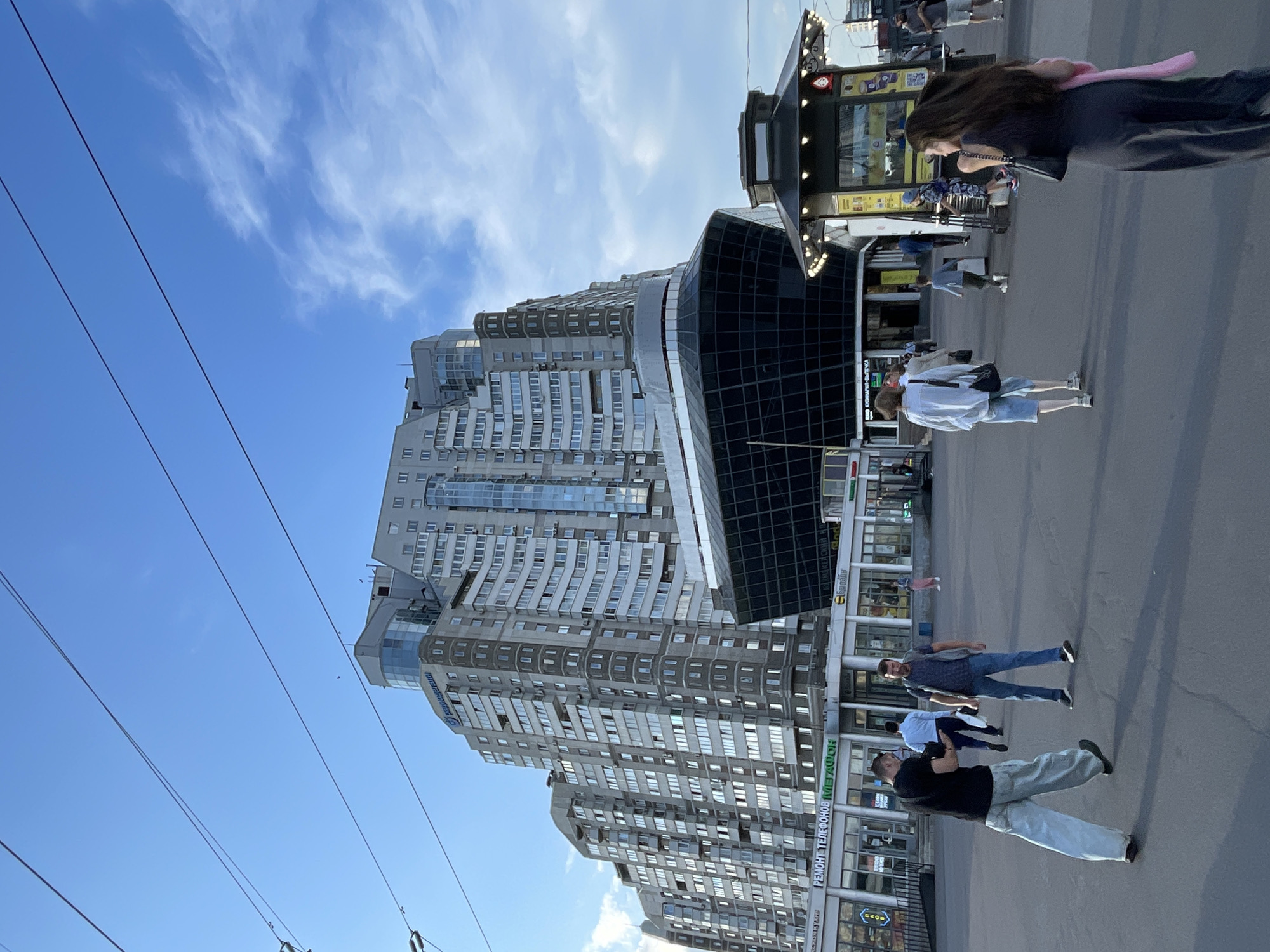 The image captures a wide-angle view of a public square in front of a modern, multi-story building complex in Saint Petersburg, Russia. The architecture of the building is striking, featuring a curved, dark-glass lower section resembling a ship's hull, topped by lighter, stepped residential levels with numerous balconies and windows. In the foreground, several people are walking and standing. Two men are engaged in conversation in the lower left, one wearing a black t-shirt and light blue pants, the other in a gray t-shirt and blue jeans. A woman in a dark dress is walking away from the camera on the right, carrying a pink scarf. Several other individuals are visible in the mid-ground, including a person in a white shirt and denim shorts walking away from the camera, and a child walking further back. Several retail establishments with signage in Russian are visible on the ground floor of the building complex. A small, ornate kiosk with lights is present on the right side of the square. The sky is blue with scattered clouds, and overhead power lines are visible, suggesting the presence of a tram or trolleybus system. The overall scene depicts an urban environment during daylight hours.