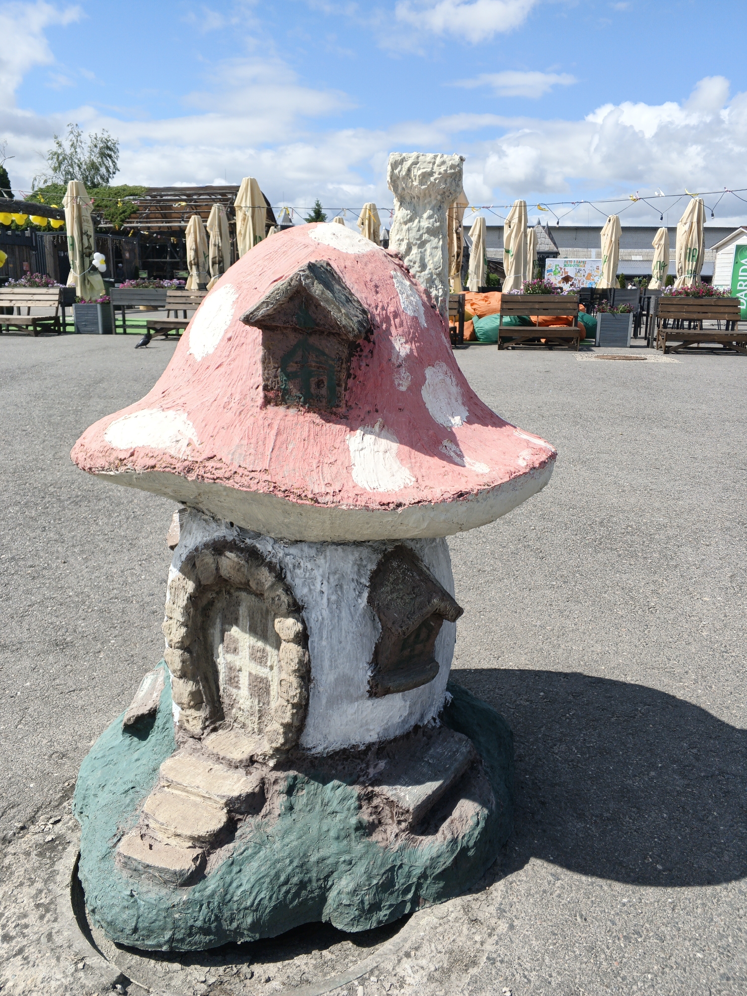 The image displays an outdoor scene on a bright, sunny day, likely at a leisure or entertainment venue in Малахавецкі сельскі Савет, Belarus. In the foreground, a prominent mushroom-shaped sculpture captures attention. Its cap is painted pink with white spots, resembling a fly agaric, and it features small, detailed windows and a round, arched door with steps, evoking a fairy-tale house. The sculpture's base is green and textured, resting on gray asphalt. Strong sunlight casts a distinct shadow of the mushroom onto the ground.
In the background, an outdoor seating or dining area is visible under a clear blue sky dotted with white clouds. Numerous light-colored, folded or partially open umbrellas are arranged, presumably to offer shade. Wooden benches and tables are set up, along with some brightly colored orange and green beanbag chairs. Planters brimming with flowers add vibrant accents. Behind these seating arrangements, various structures can be seen, including a rustic wooden building with a vine-covered roof and another building further back. A tall, craggy, light-colored sculpture or monument stands to the left in the background.
A small, dark bird, possibly a crow or pigeon, can be observed on the paved ground in the middle distance. The scene suggests a relaxed atmosphere in an area designed for recreation.
Visible text includes "BARIDA" on a green sign to the right of the seating area. A large white sign or poster in the background features colorful illustrations and Cyrillic text, partially decipherable as "РАЗВЛЕЧЕНИЯ" (Entertainment) and "ДЛЯ МАЛЕНЬКИХ ОТДЫХАЮЩИХ" (for little vacationers/guests), indicating a family-friendly establishment or children's activity zone.