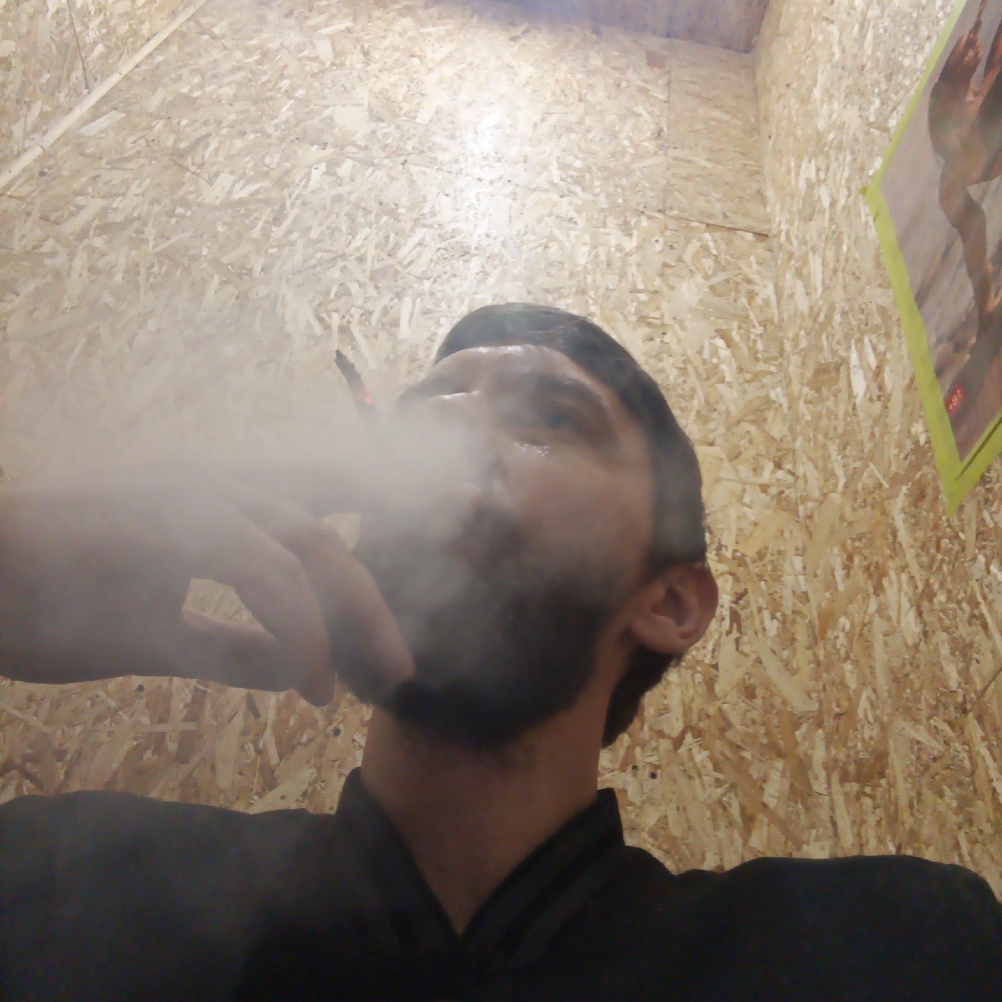 A man with a dark beard is smoking a cigarette, producing a cloud of smoke. He is wearing a black shirt. The background is a wall made of particleboard. A poster hangs on the wall. The photo was taken at a low angle, looking up at the man. The location is Chaikovsky, Russia.