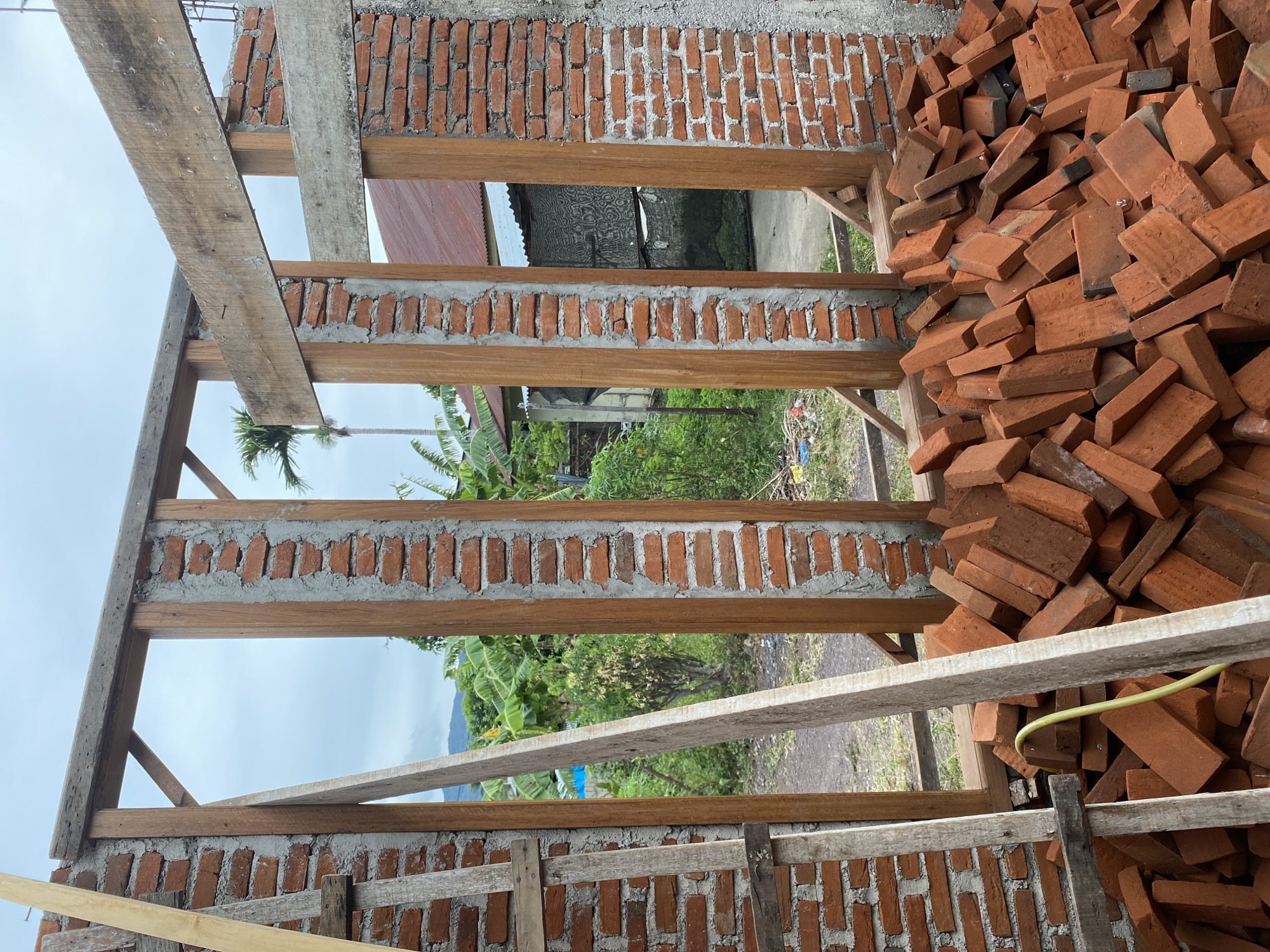 The image depicts the construction of a building in Banda Aceh, Indonesia. The scene shows a partially built brick wall with wooden framing, likely for a window or doorway. A large pile of red bricks is in the foreground, ready for use. Beyond the construction, lush green vegetation, including banana trees and a palm tree, is visible, characteristic of the tropical climate of Banda Aceh. The sky is overcast. There are no people visible in the image.