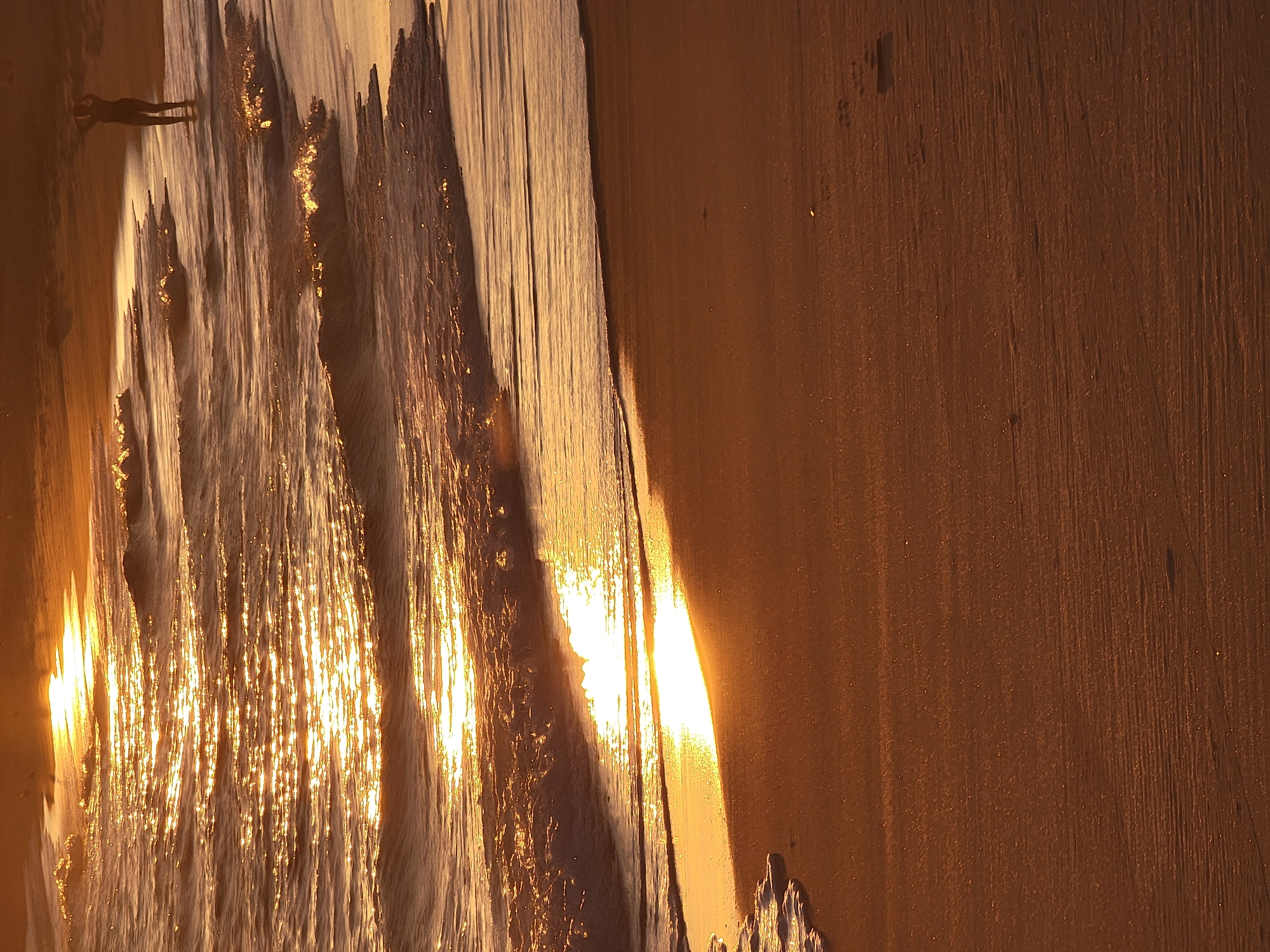 A person stands in shallow water at the edge of a beach at sunset. The sun's reflection creates a golden shimmer on the water's surface. Waves gently lap at the shore, and the sand is a warm, sandy brown. The scene is likely Quarteira, Portugal, given the description. The person, silhouetted against the light, has their hands raised as if adjusting their hair. The overall mood is tranquil and serene, with the natural beauty of the sunset and the ocean taking center stage.