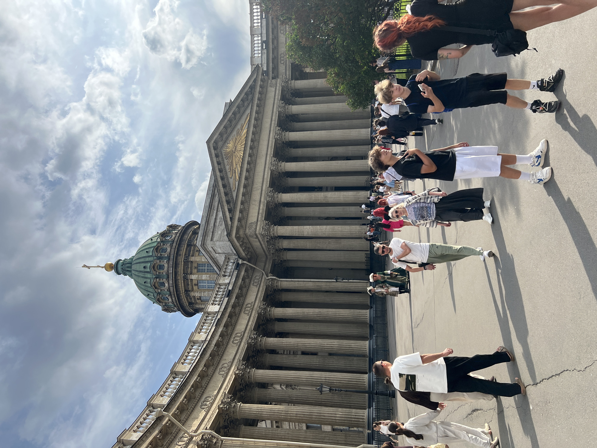 The image depicts a sunny day in Saint Petersburg, Russia, likely during the daytime, with a bright sky and scattered clouds. The scene features a large, classical building with a prominent green dome and a colonnade of imposing stone columns. A crowd of people is gathered in front of the building, some walking and others standing.
The people in the foreground are dressed in casual, modern attire. Several young individuals are visible, with one person wearing a black t-shirt and black shorts, holding a phone. Another person is in a black t-shirt and white shorts, with white sneakers. Further back, a person with red-dyed hair is visible in the upper part of the image, wearing a black dress. The diversity of clothing suggests a mix of locals and tourists.
The architectural style of the building, with its dome and grand columns, is characteristic of Saint Petersburg, hinting at a significant landmark such as a cathedral or museum. The activity appears to be a casual gathering, possibly for sightseeing or attending an event. The overall mood seems relaxed and observational, with people taking in the surroundings. No visible text is present in the image.