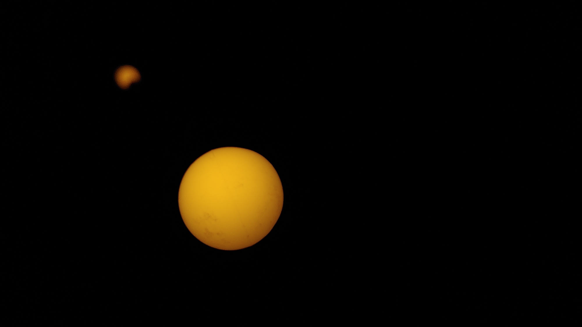 The image shows a celestial event with two bright, yellow-orange spheres against a black background. The larger sphere is on the left side of the frame and appears to be partially obscured from the bottom. The smaller sphere is in the upper right quadrant, and a portion of it appears to be covered by a darker, crescent shape, suggesting an eclipse or transit. There are no discernible details of the city of Mukachevo, Ukraine, or any human presence in the image. The focus is solely on the astronomical phenomena.