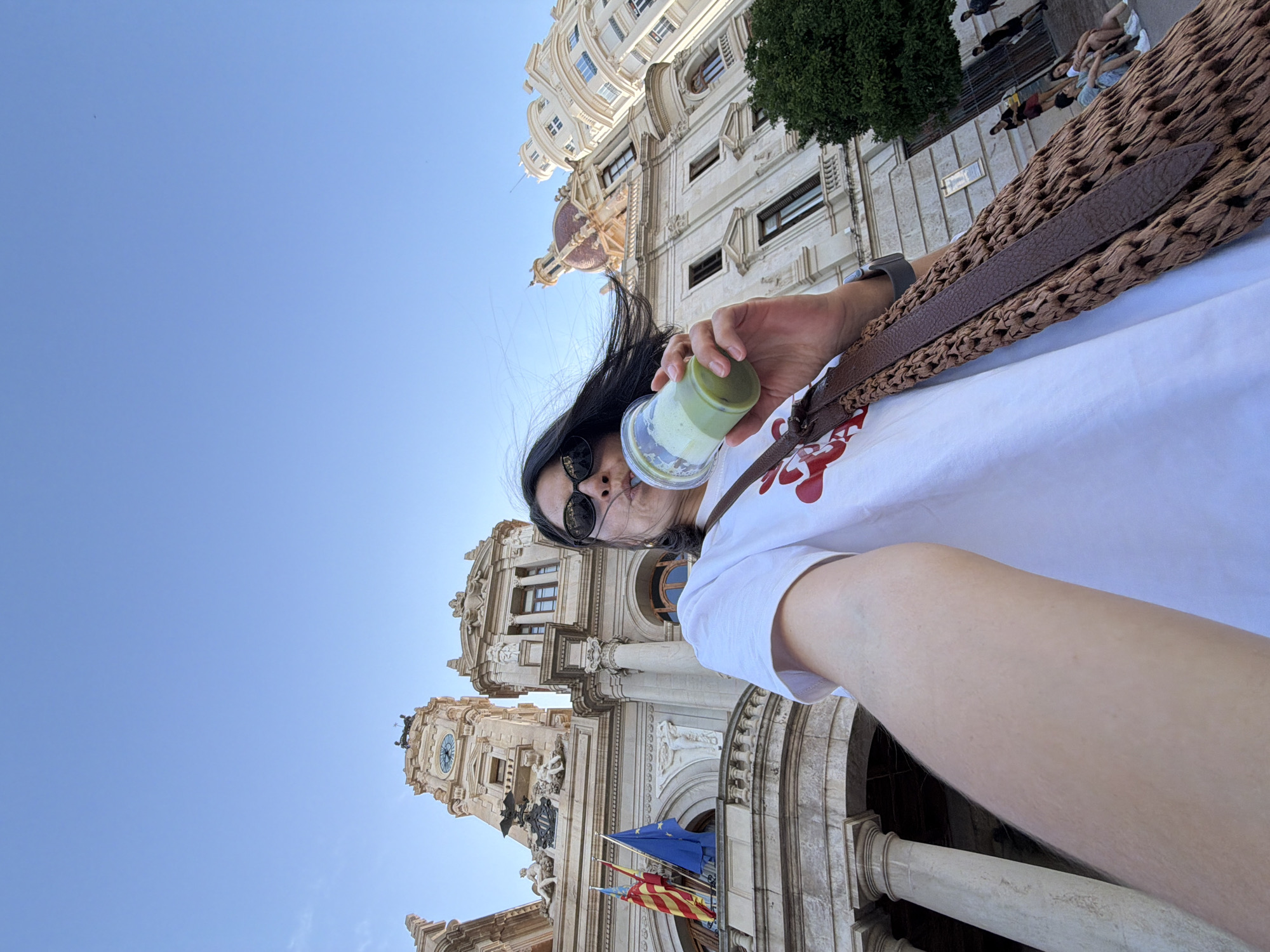 This is a wide-angle, low-angle selfie taken outdoors in front of the Valencia City Hall in Valencia, Spain. A person is in the foreground, holding a clear plastic cup with a green beverage and a straw to their mouth. They are wearing a white t-shirt with a red design, sunglasses, and have dark hair being blown by the wind. A woven brown bag with leather straps is slung across their shoulder.
The background features the ornate facade of the Valencia City Hall, characterized by classical architecture, columns, balconies, flags (including the Valencian flag and the European Union flag), and a prominent clock tower. To the right of the City Hall, another cream-colored building with distinctive dome-like structures is visible. The sky is clear and blue. In the distance, several people are sitting and standing on a paved area, some appearing to be tourists. A green tree is also visible on the right side of the frame. The lighting suggests it is daytime, likely late afternoon due to the angle of the light and the long shadows.