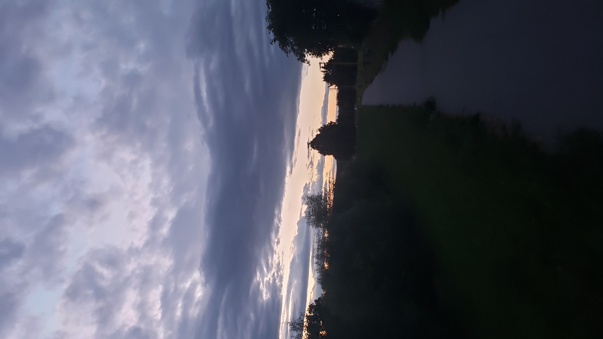 The image depicts a twilight scene with a dramatic sky filled with dark, textured clouds interspersed with lighter patches where the sun is setting. The horizon line is low, allowing the sky to dominate the frame. In the foreground, a paved pathway extends into the distance, bordered by grassy areas and dark silhouettes of trees and bushes on either side. The overall impression is one of a peaceful, possibly rural or suburban, evening. Without further information or identifying landmarks, it is impossible to pinpoint the exact location within the city of Prešov, Slovakia.