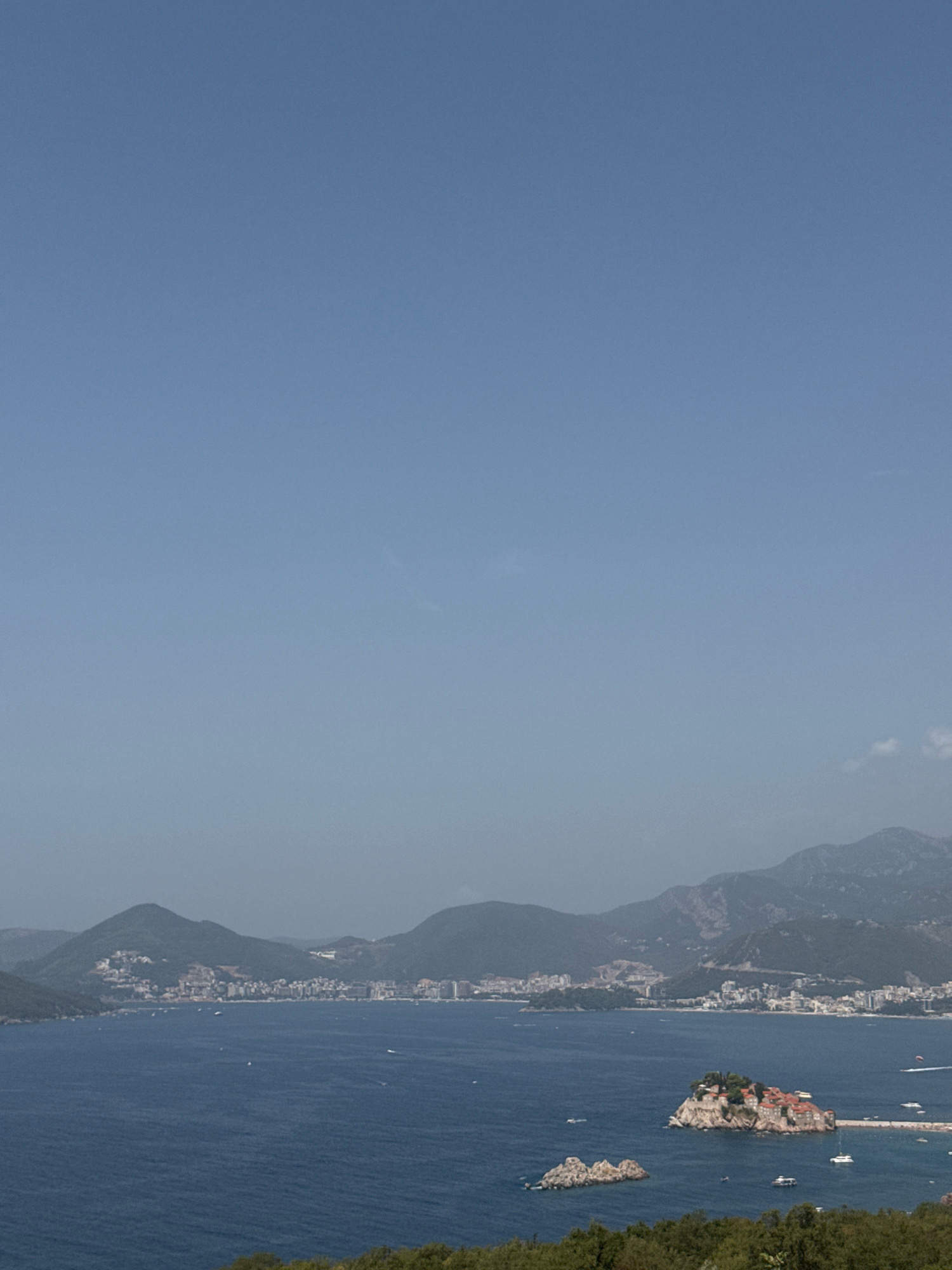An aerial, wide shot captures the mountainous coastline of Vrba, Montenegro, under a clear, pale blue sky. The dark blue sea occupies the lower half of the frame, dotted with a few boats and a small islet with buildings and trees connected to the mainland by a causeway. The coastline is lined with a sprawling town and more mountains rise behind it. The foreground features a dense green forest.