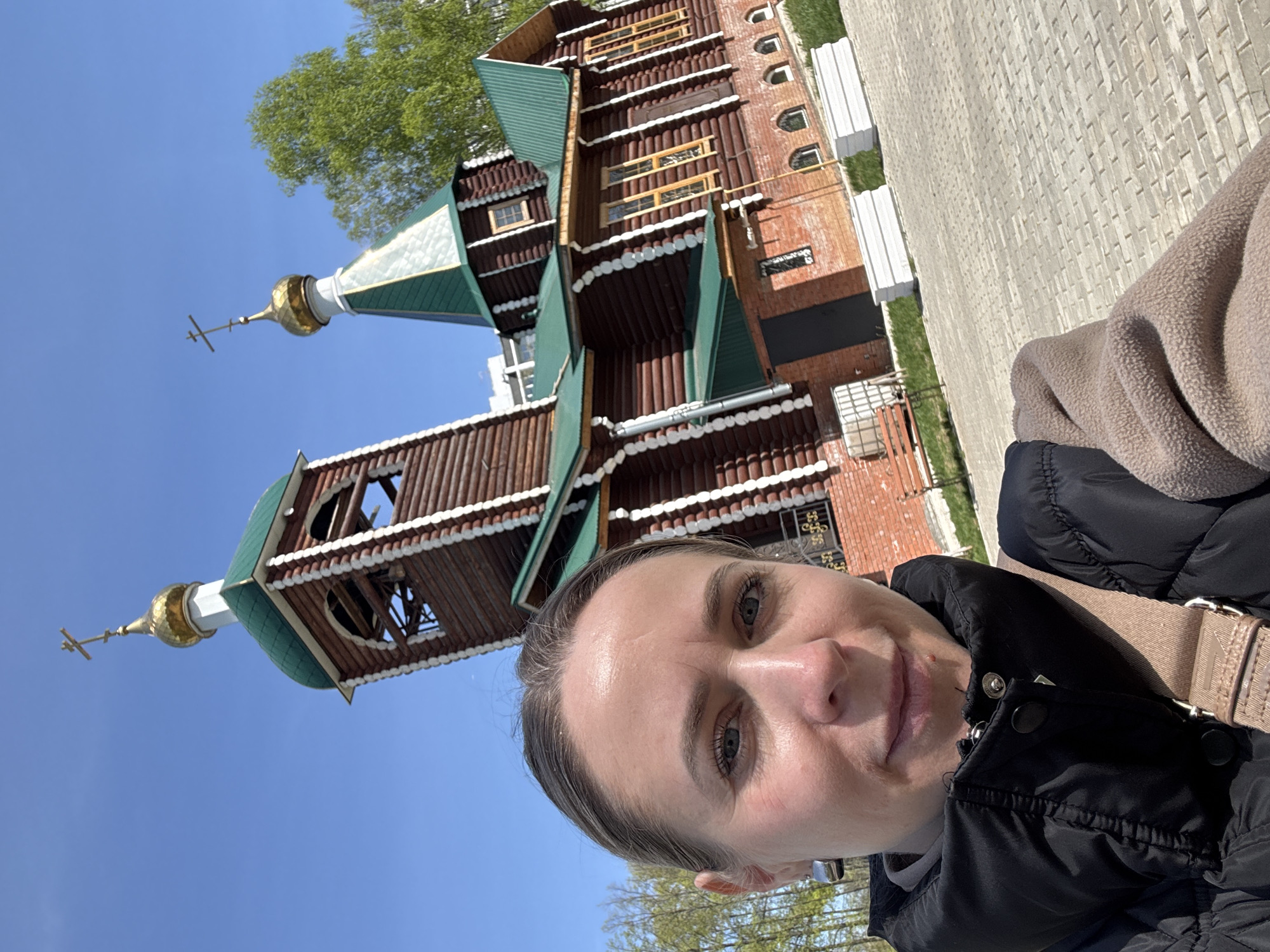 A woman is taking a selfie outdoors, with a Russian Orthodox church in the background. The church is made of brown logs and red brick, with green roofs and gold domes topped with Orthodox crosses. It has an ornate, traditional design. The sky is blue, and some trees with green leaves are visible. The woman is wearing a black jacket and a beige shoulder bag. The location is Chaikovsky, Russia.